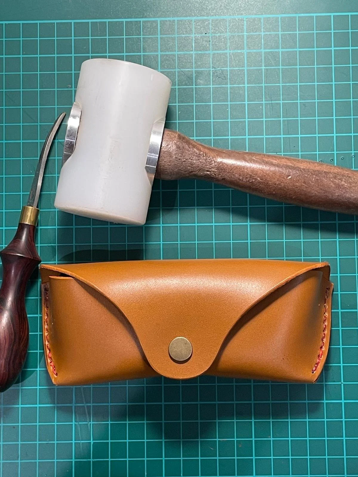A leather tool pouch, a wooden mallet with a white rubber head, and a carving tool with a wooden handle are placed on a green cutting mat.