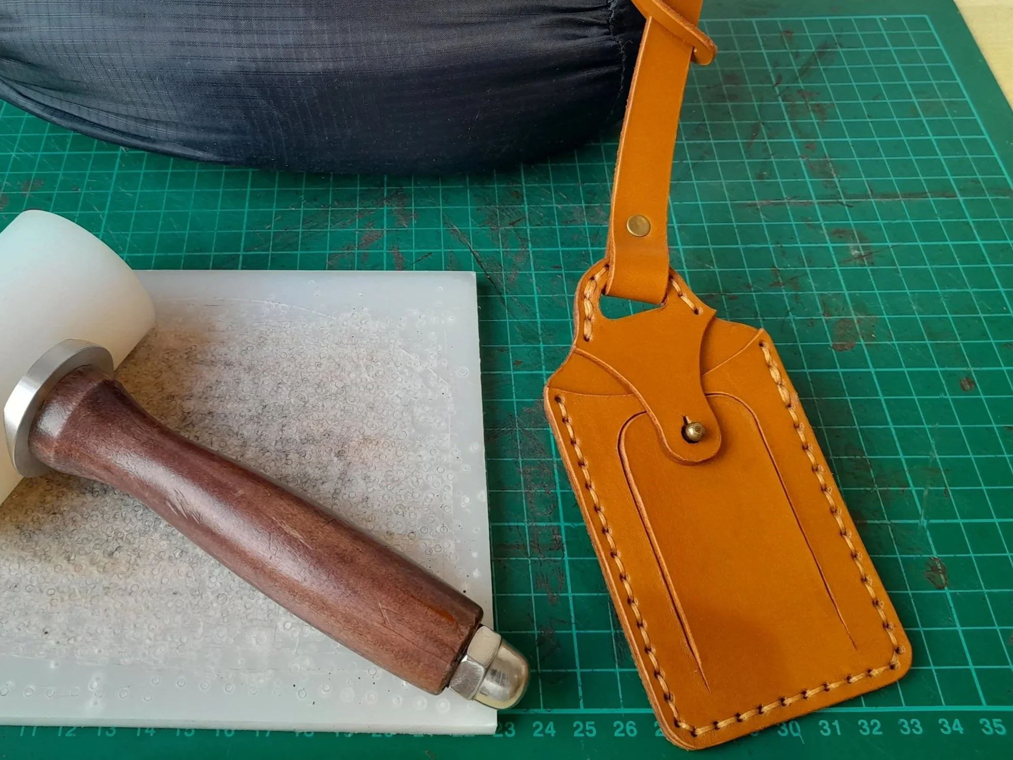 Craft tools including a leather mallet, green cutting mat, black carrying case, and orange leather luggage tag on a worktable.