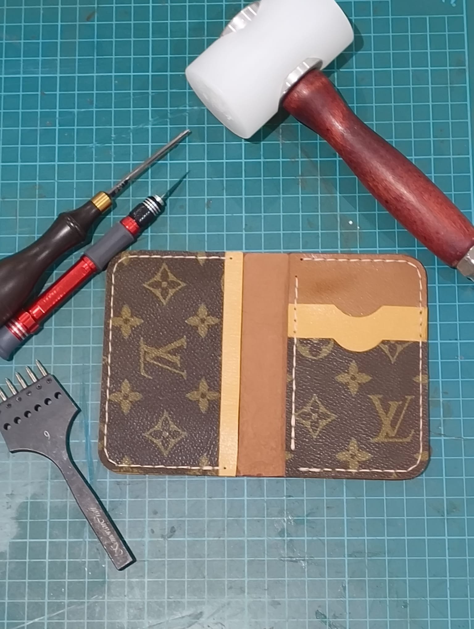 A green cutting mat with a Louis Vuitton wallet, two screwdrivers, a metal ruler, and a material piece.