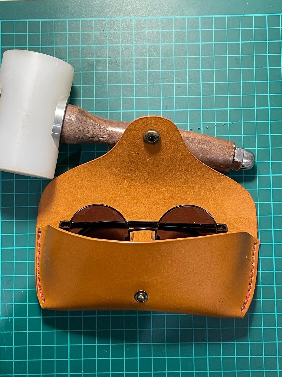 A pair of round black sunglasses inside a tan leather case, with a wooden hammer and a white object on a green grid cutting mat.