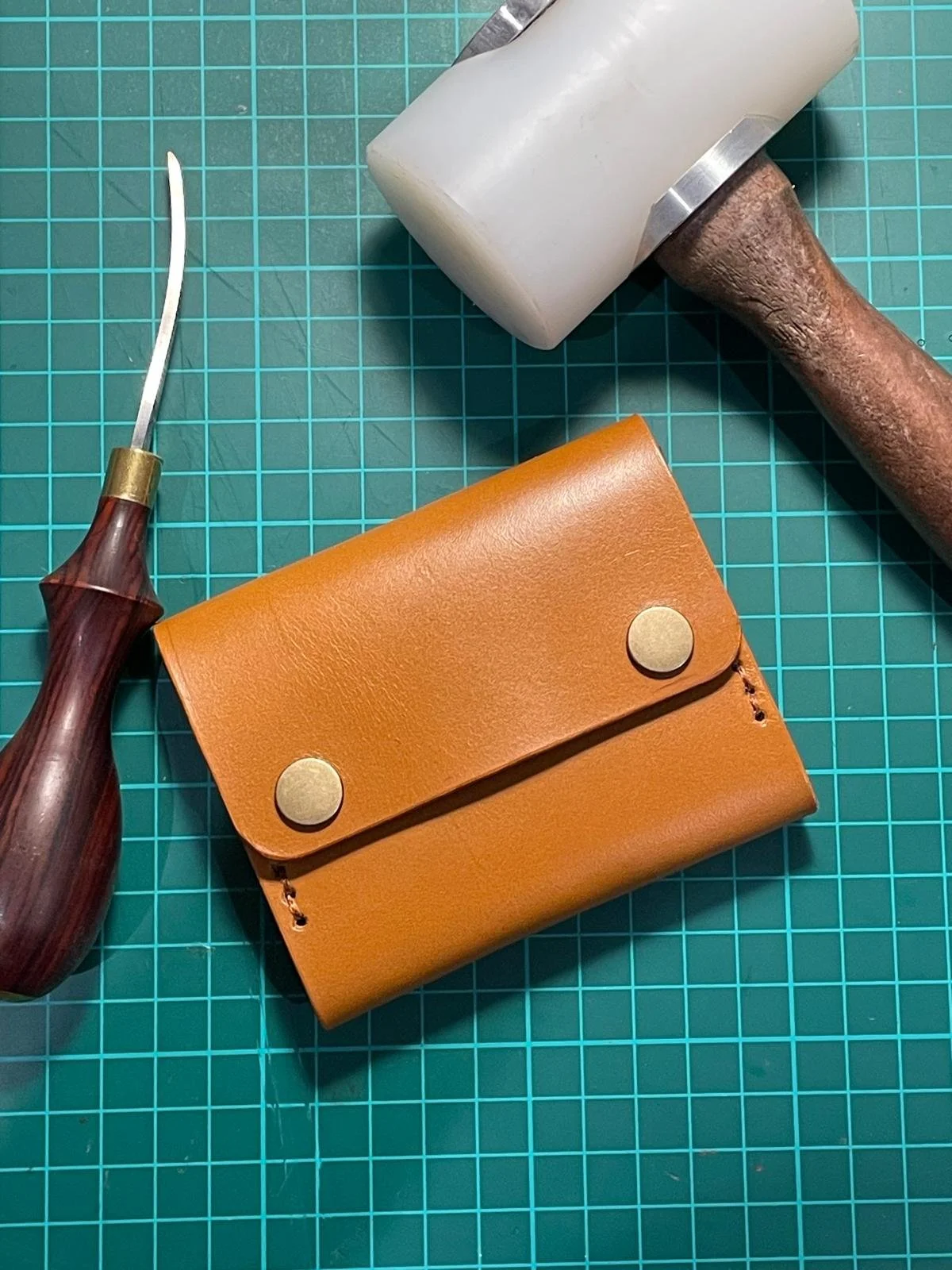A leather wallet, a mallet, a small carving tool, and a cutting mat with grid lines.