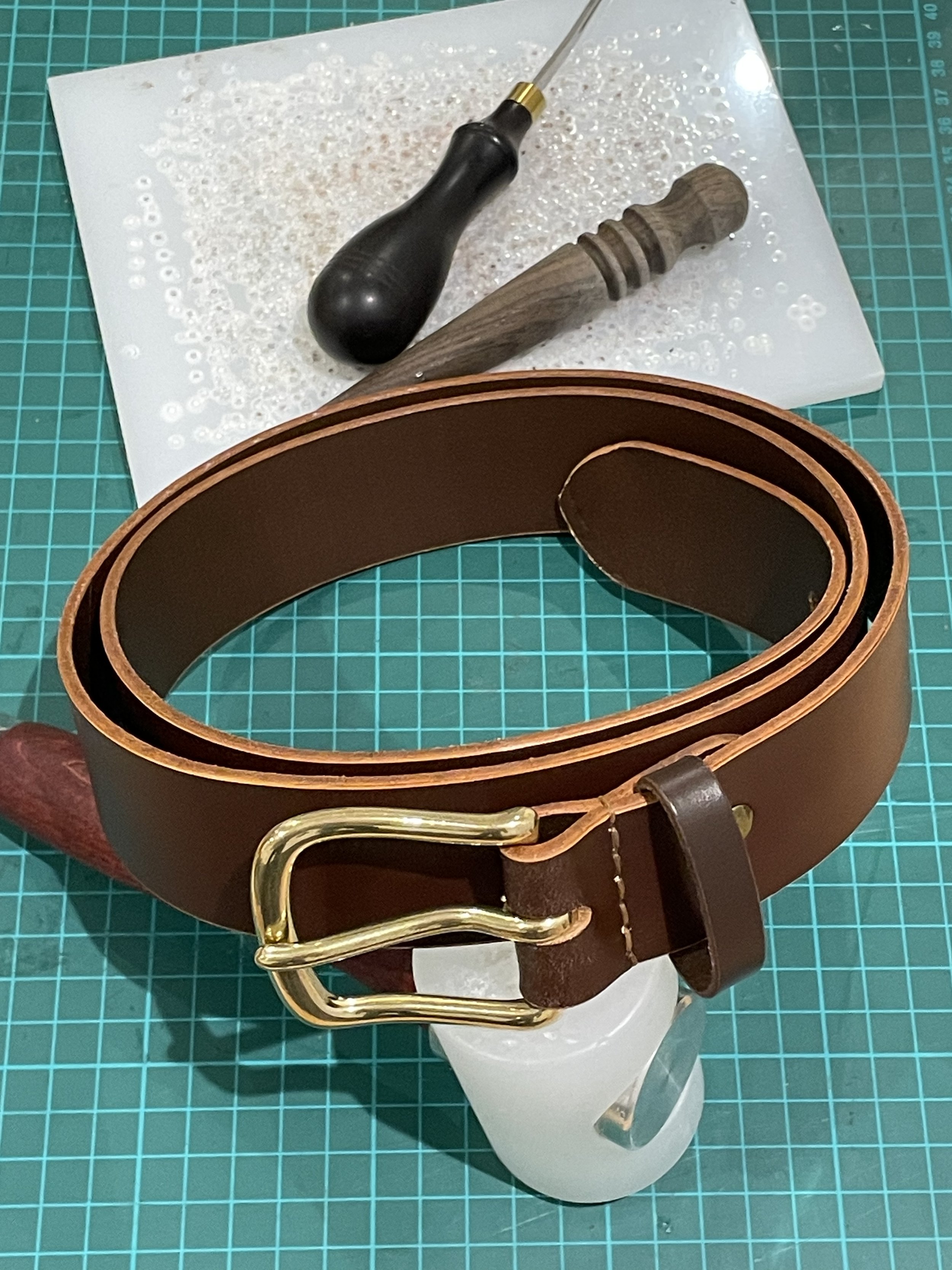 A leather belt with a gold buckle, a wooden and metal belt roller, and a screwdriver, placed on a cutting mat with a white paper underneath.
