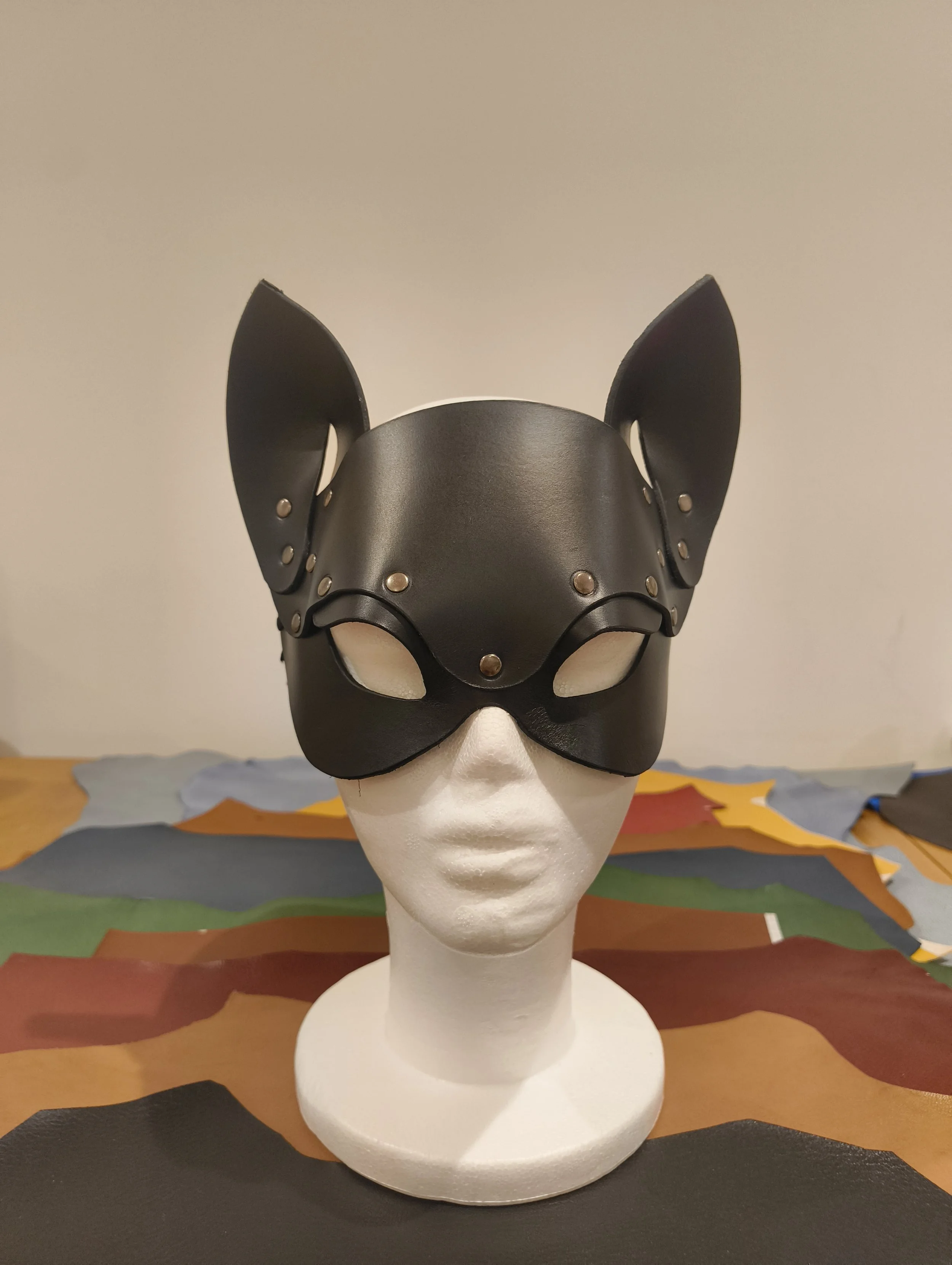 A mannequin head displays a black leather mask with cat-like ears and metallic rivets, set against a plain wall background.