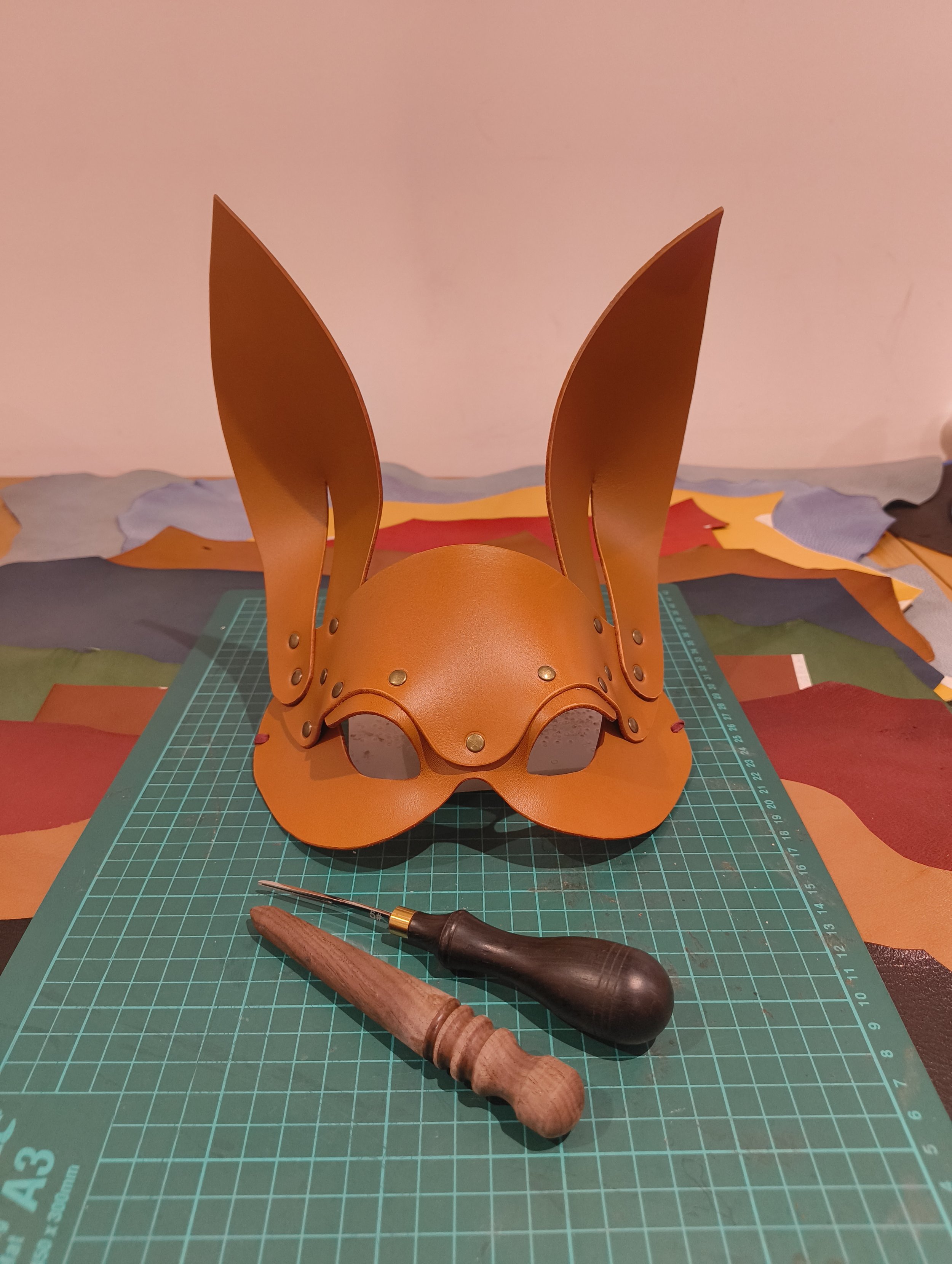 A leather headpiece with large, pointed ears, placed on a green cutting mat, with precision tools nearby. The background features color swatches and craft supplies.