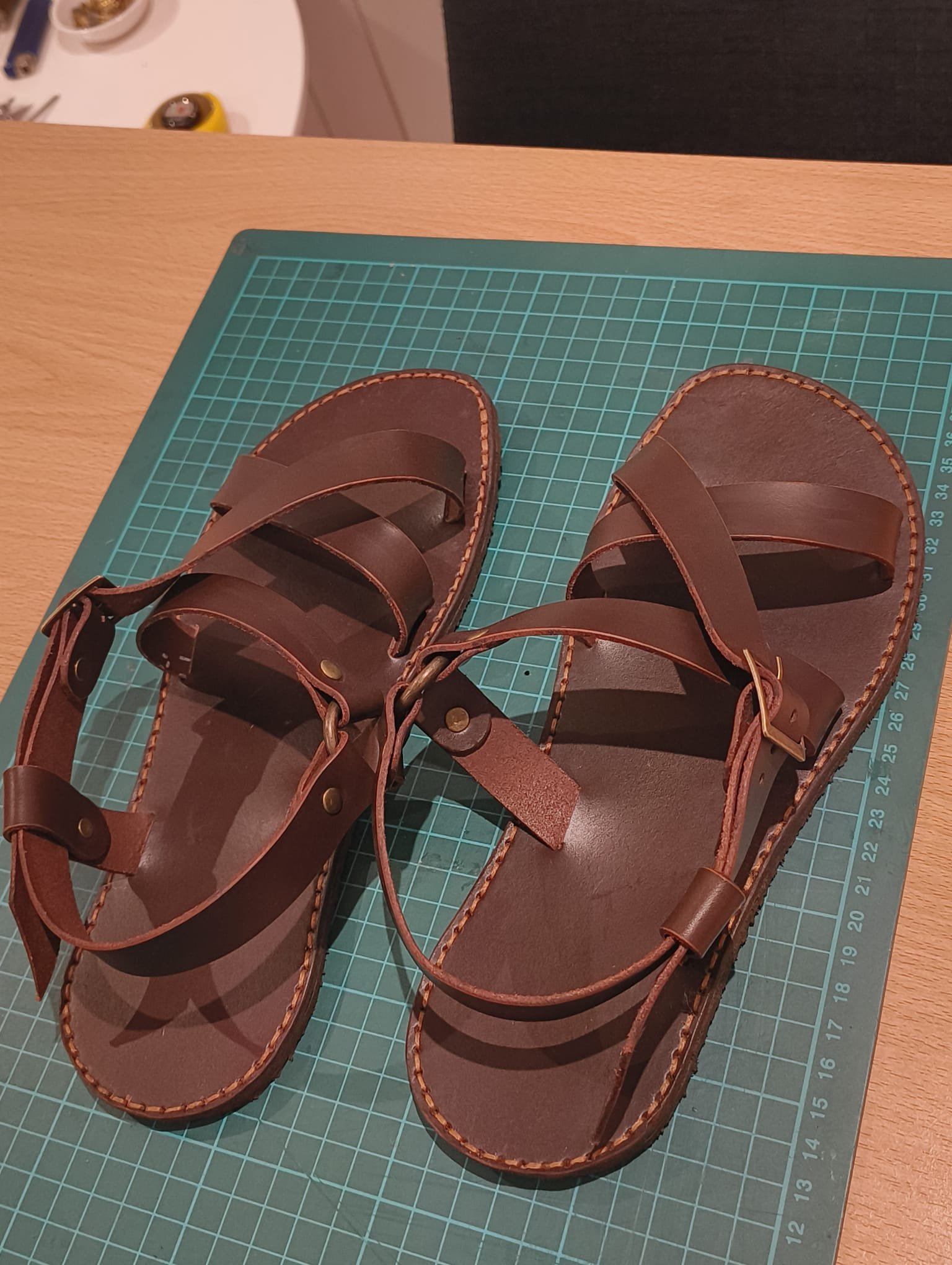 A pair of brown leather sandals with multiple straps and stitching detail, placed on a green cutting mat on a wooden surface.