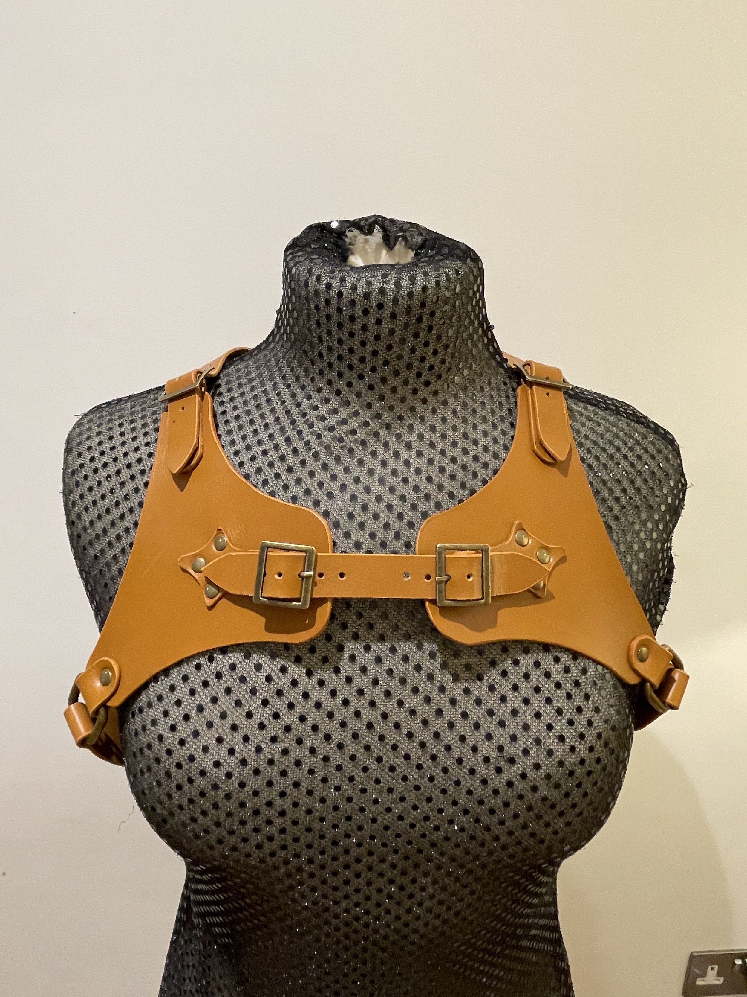 A black mesh mannequin torso wearing an orange harness with metal buckle details, set against a plain light-colored wall.