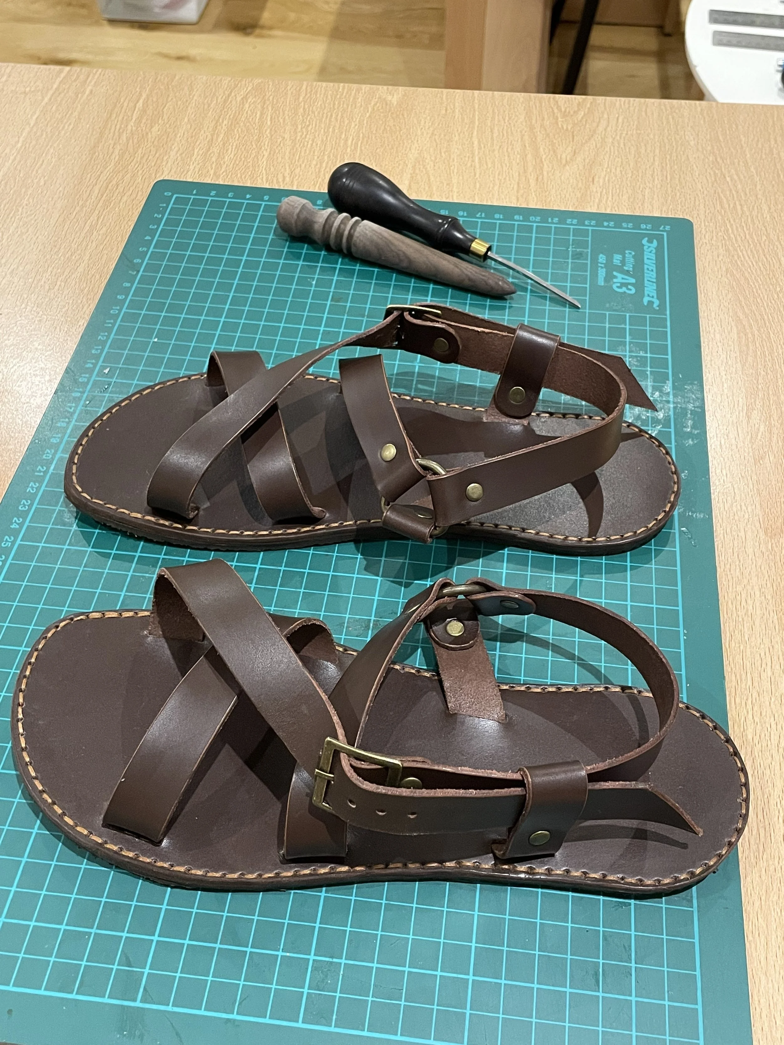 A pair of brown leather sandals with adjustable straps and brass buckles, placed on a green cutting mat on a wooden table, with tools including a small screwdriver and a wooden-handled awl in the background.