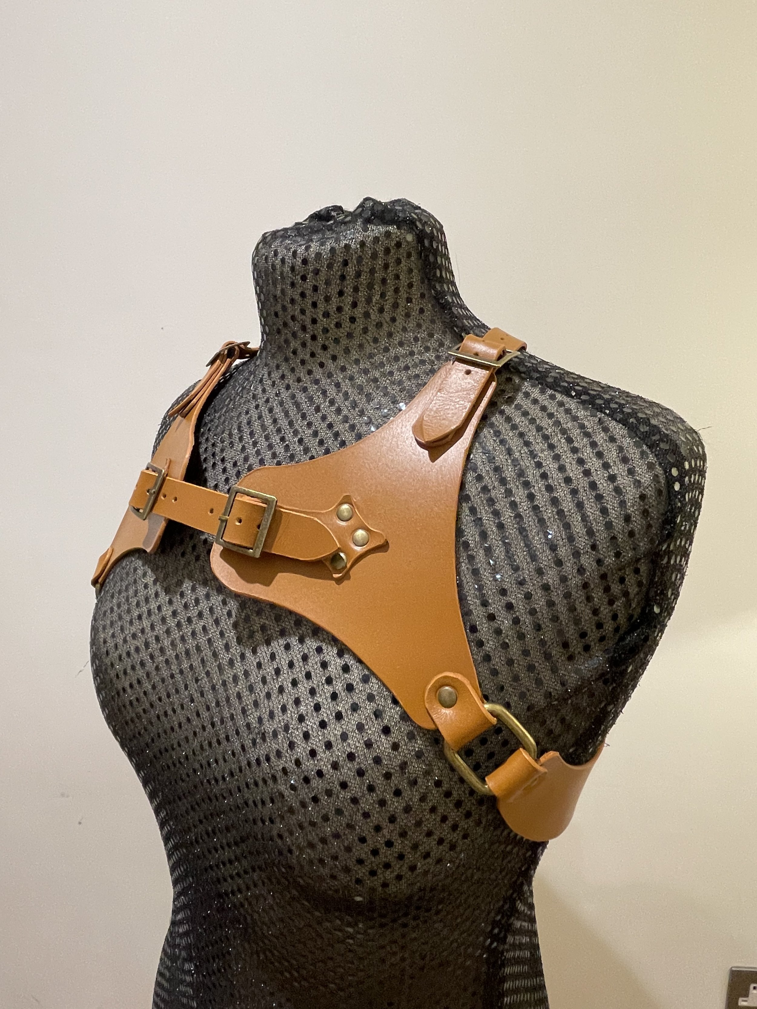 A display mannequin's upper body wearing a tan leather harness with metal buckle and rivets, set against a plain off-white background.