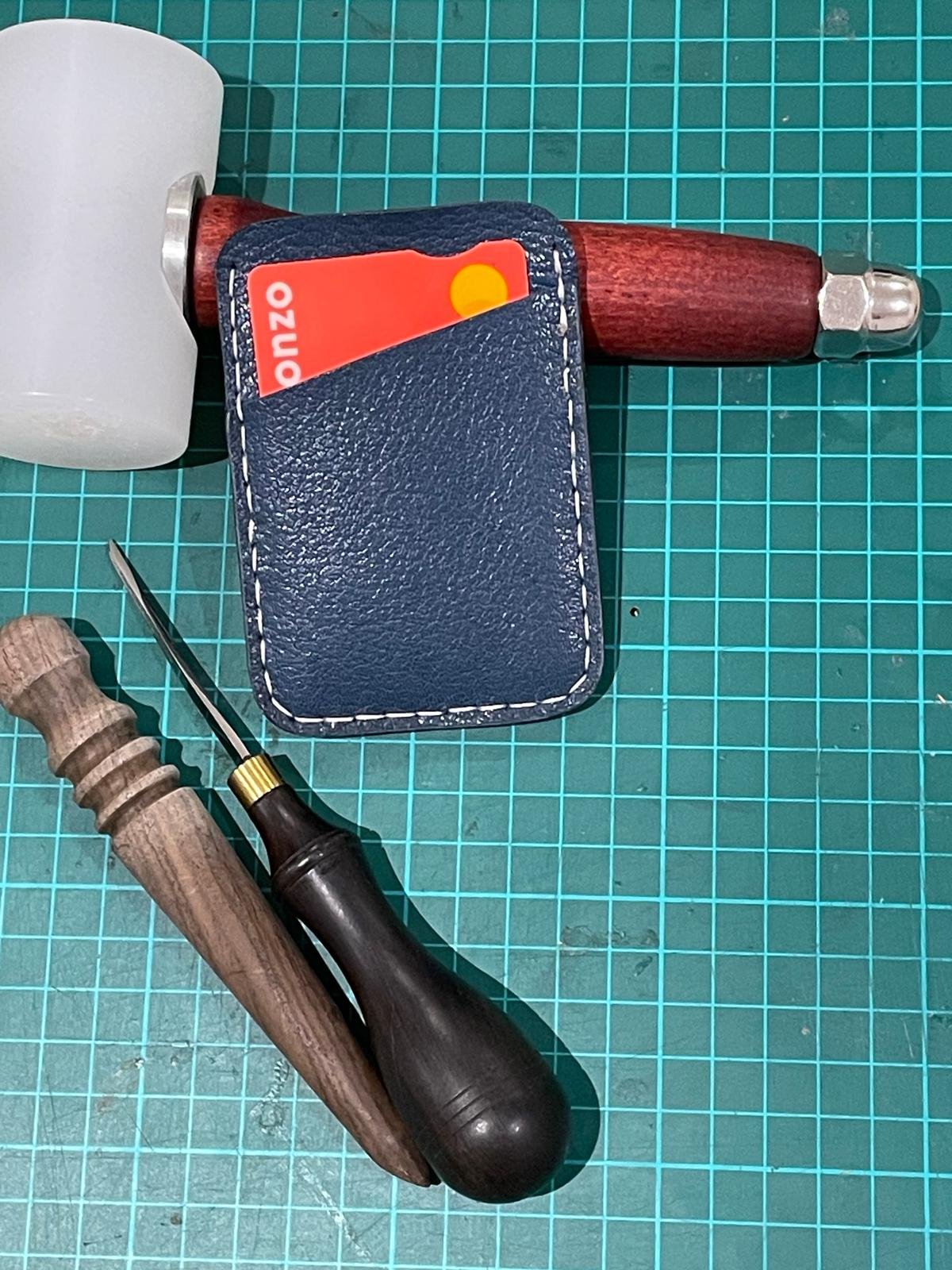 A green cutting mat with a rubber mallet, a leather wallet with a credit card, a small flathead screwdriver, and a wood-handled tool.
