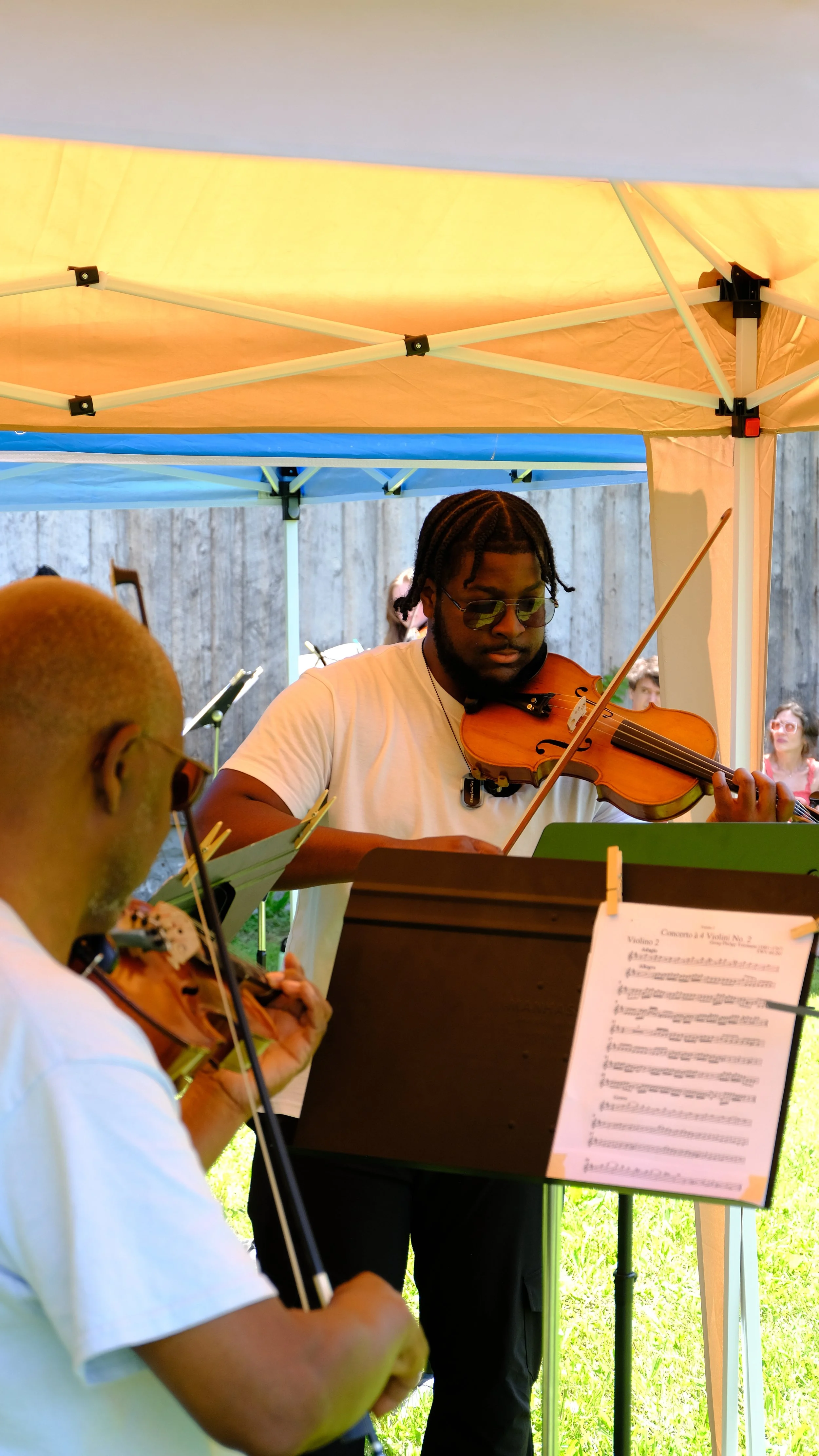 Two musicians playing violins under a canopy during an outdoor event, with a wooden fence and some people sitting in the background.
