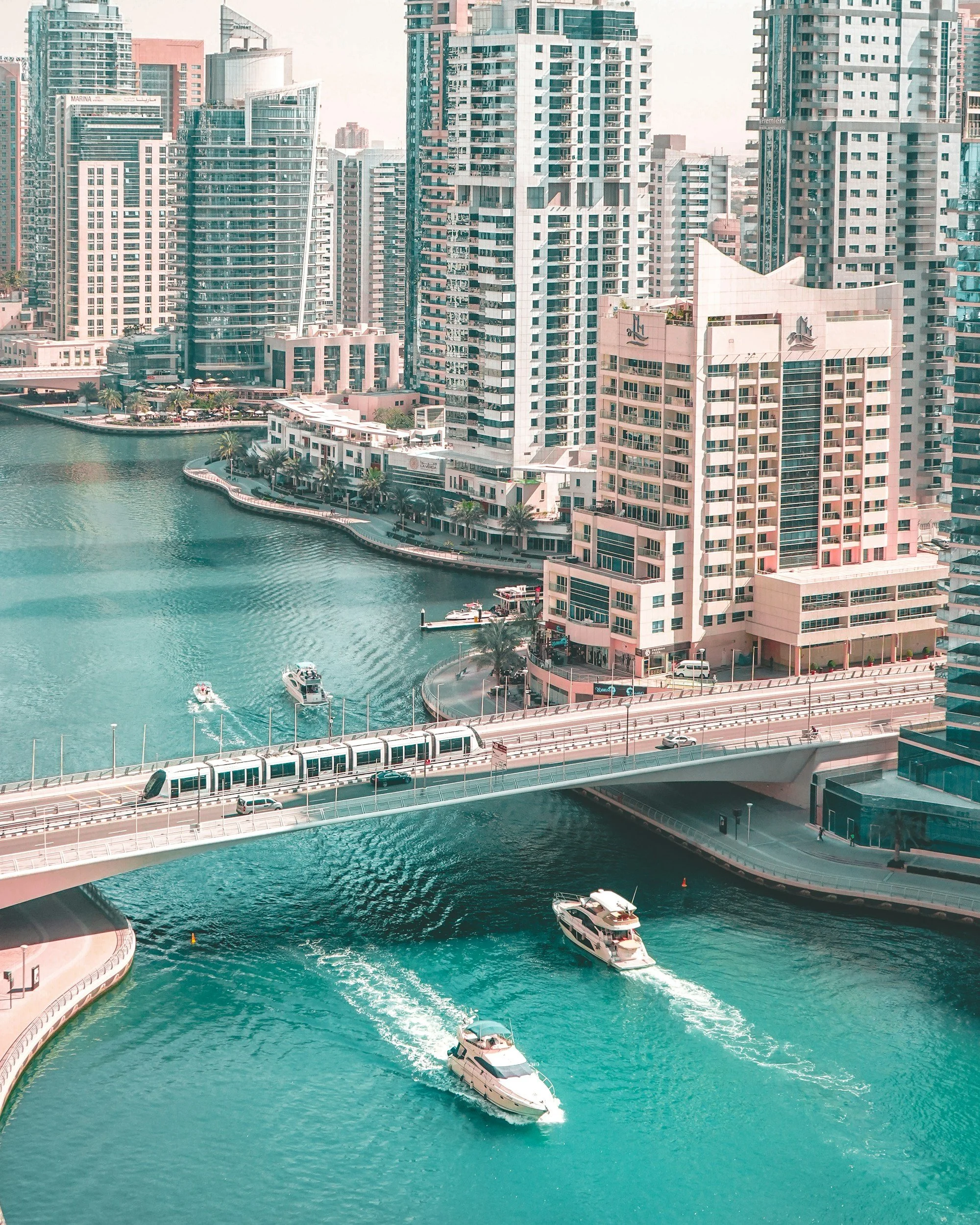 Skyline of modern high-rise buildings along a waterway with boats, a bridge with a moving train, and a boat cruising in the water.