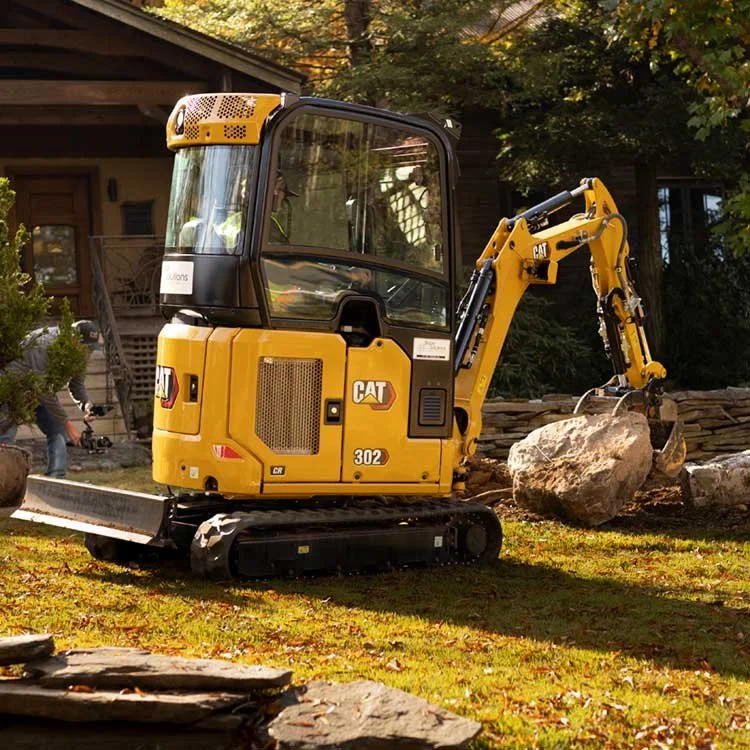 A small yellow CAT mini excavator moving a large rock in a backyard with trees and a house in the background.