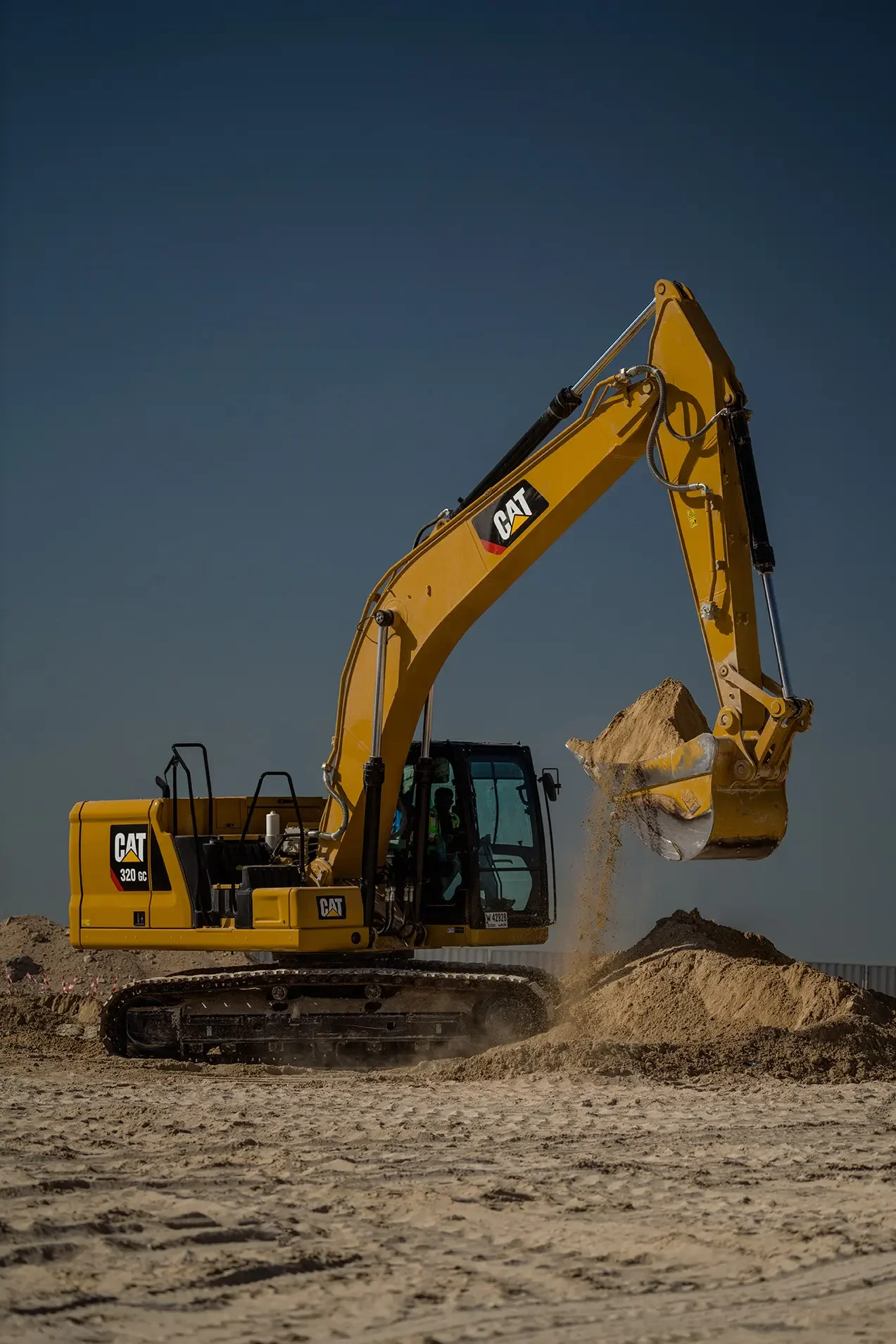 A yellow Caterpillar 320 GC excavator lifting and pouring dirt onto a mound at a construction site under a clear blue sky.