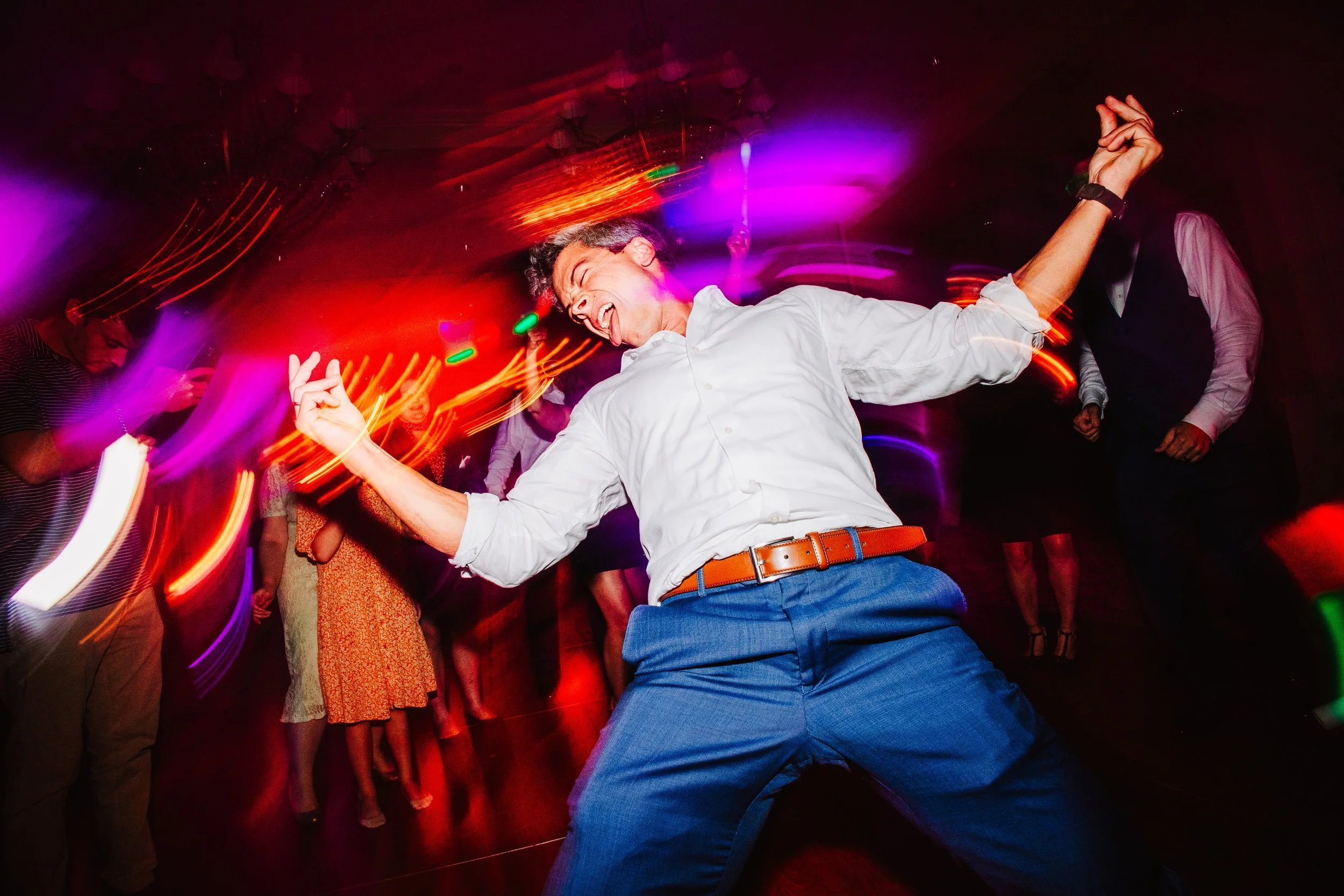 A man dancing energetically on a dance floor at a party, with colorful lights and other people in the background.