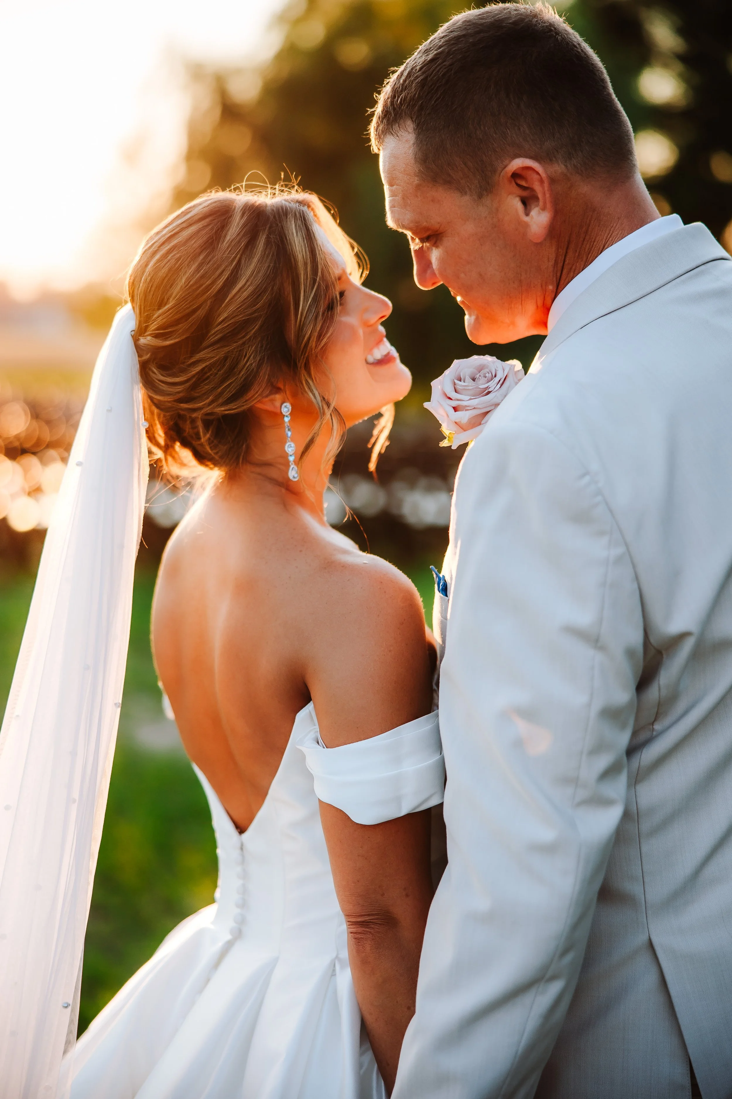 A bride and groom share a romantic moment outdoors during sunset, smiling and gazing into each other's eyes.