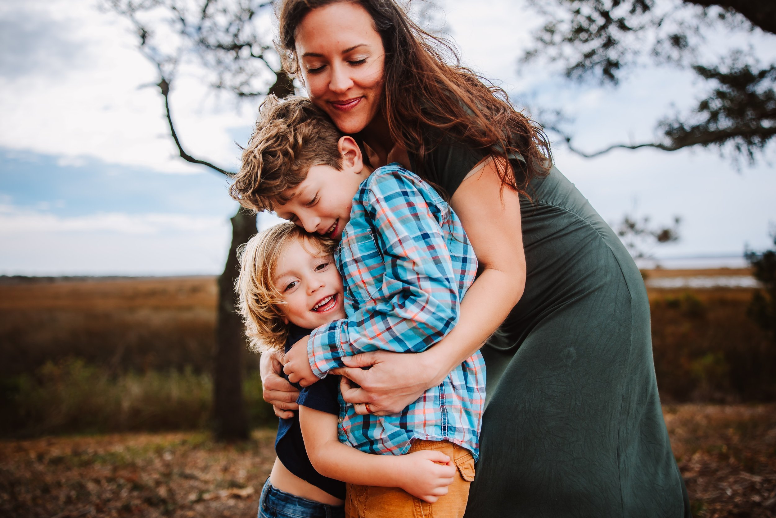 A woman hugging two children outdoors, with trees and a cloudy sky in the background.