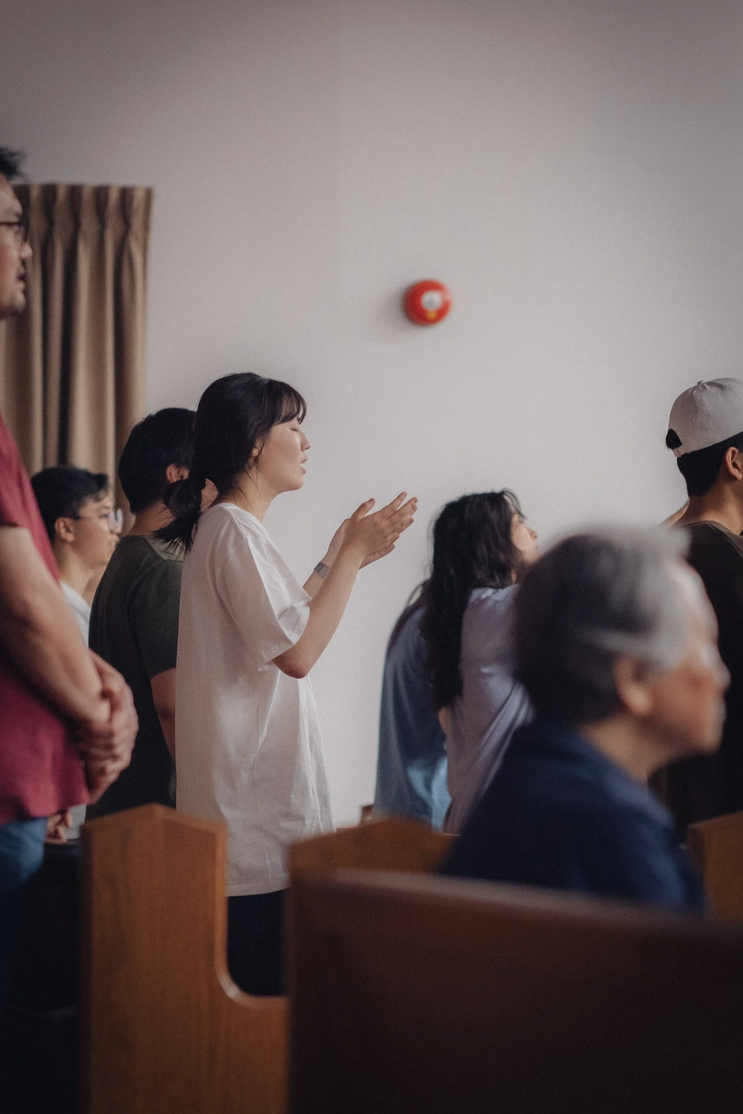 A group of people, mostly women, are standing and seated in a room, with some clapping and others praying or meditating, inside a room with plain white walls and beige curtains.