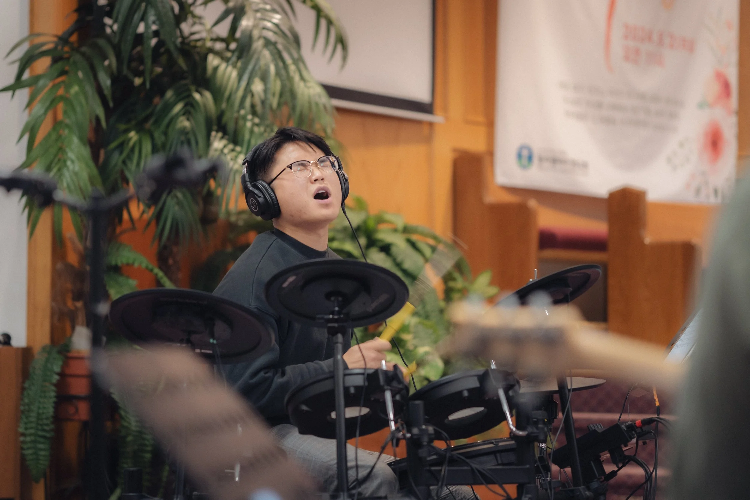 A young man wearing glasses and headphones playing electronic drum set inside a room with wooden walls and green plants.