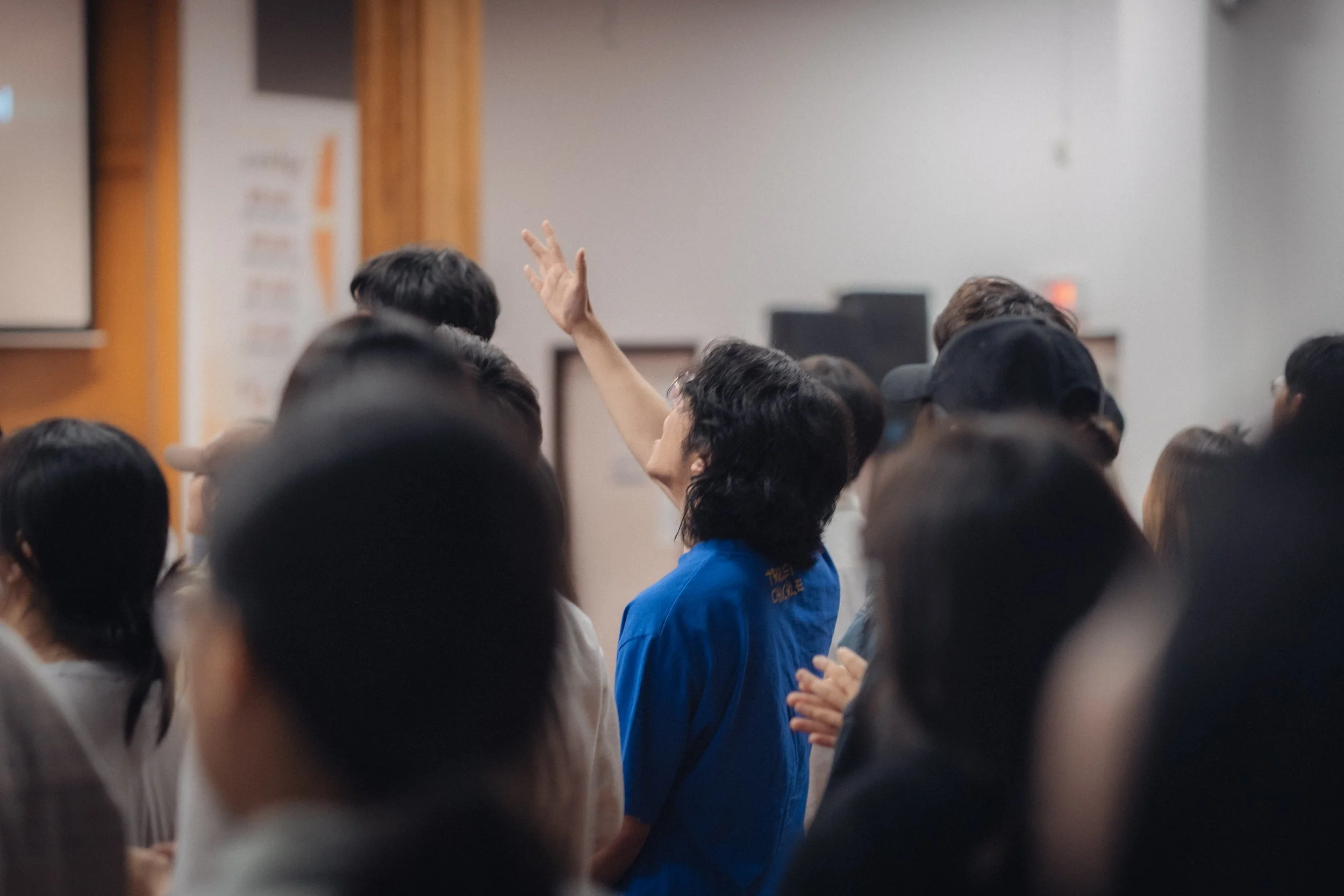 A woman in a blue shirt raises her hand in a crowded room during a presentation or meeting.