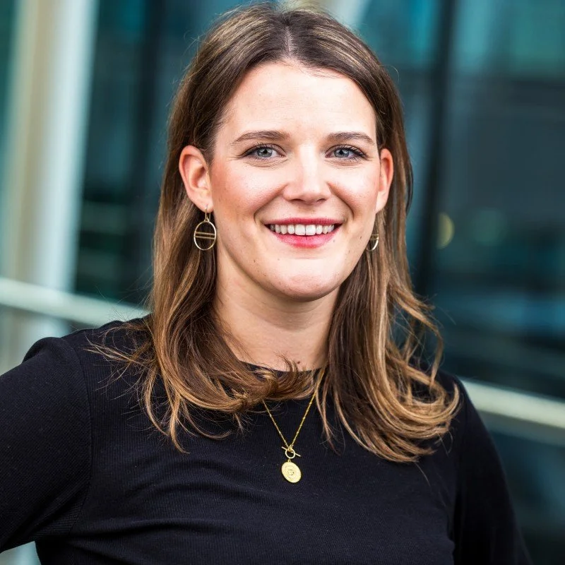 Porträt einer lächelnden jungen Frau mit braunen Haaren, schwarzen Oberteil, Schmuck im Hintergrund eines modernen Gebäudes.