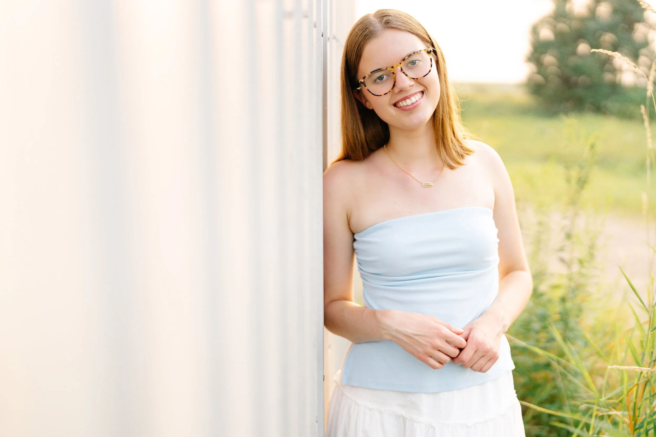 Minnesota senior portrait photographer, LKW Photography, photographs a high school senior with glasses, and a white strapless top, smiling and standing outdoors next to a white wall, with greenery in the background.
