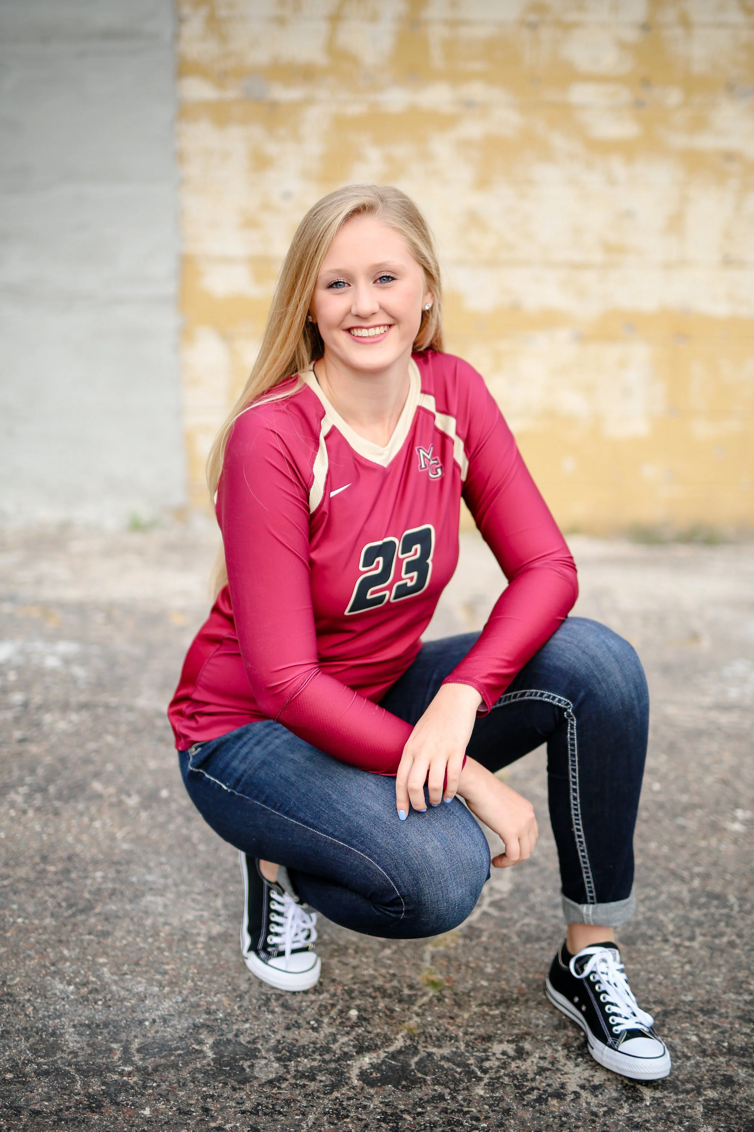 Minnesota senior photographer, LKW Photography, photographs a young woman with blonde hair, wearing a maroon sports jersey with the number 23, dark jeans, and black sneakers, squatting on an asphalt surface in front of a weathered yellow brick wall.