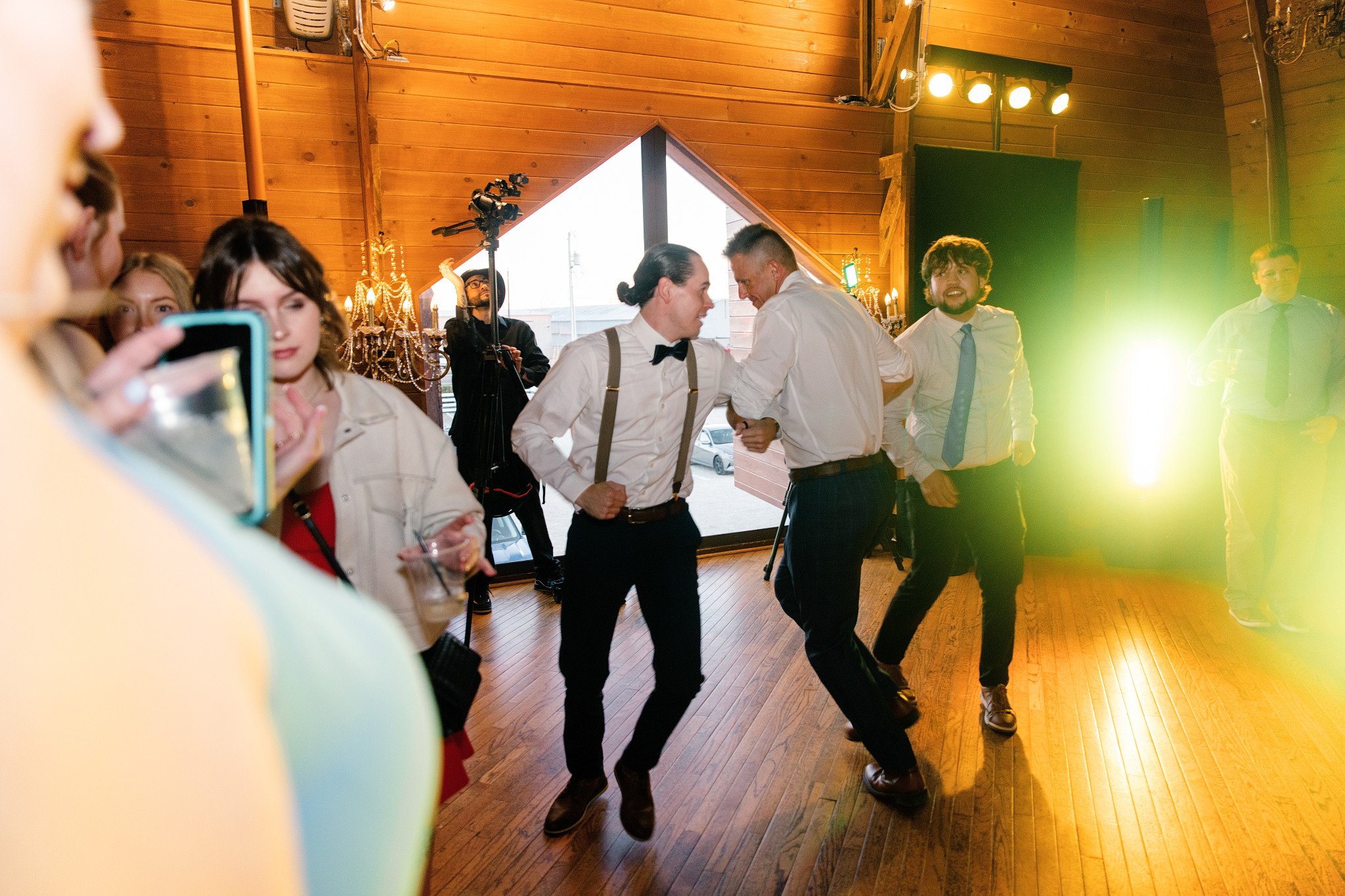 People dancing and socializing inside a wooden venue with warm lighting and chandeliers.