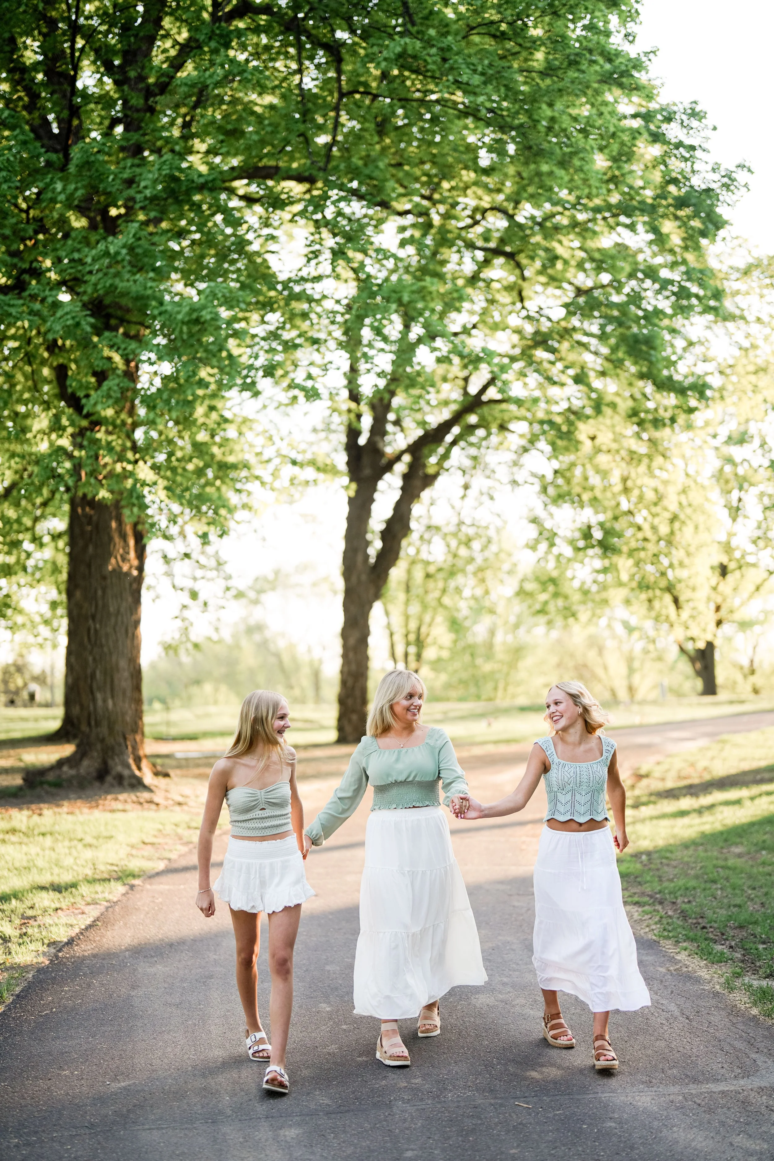 Three women, two young girls and an older woman, walking hand-in-hand on a paved path in a park with green trees and sunlight, taken by Minnesota family photographer, LKW Photography.