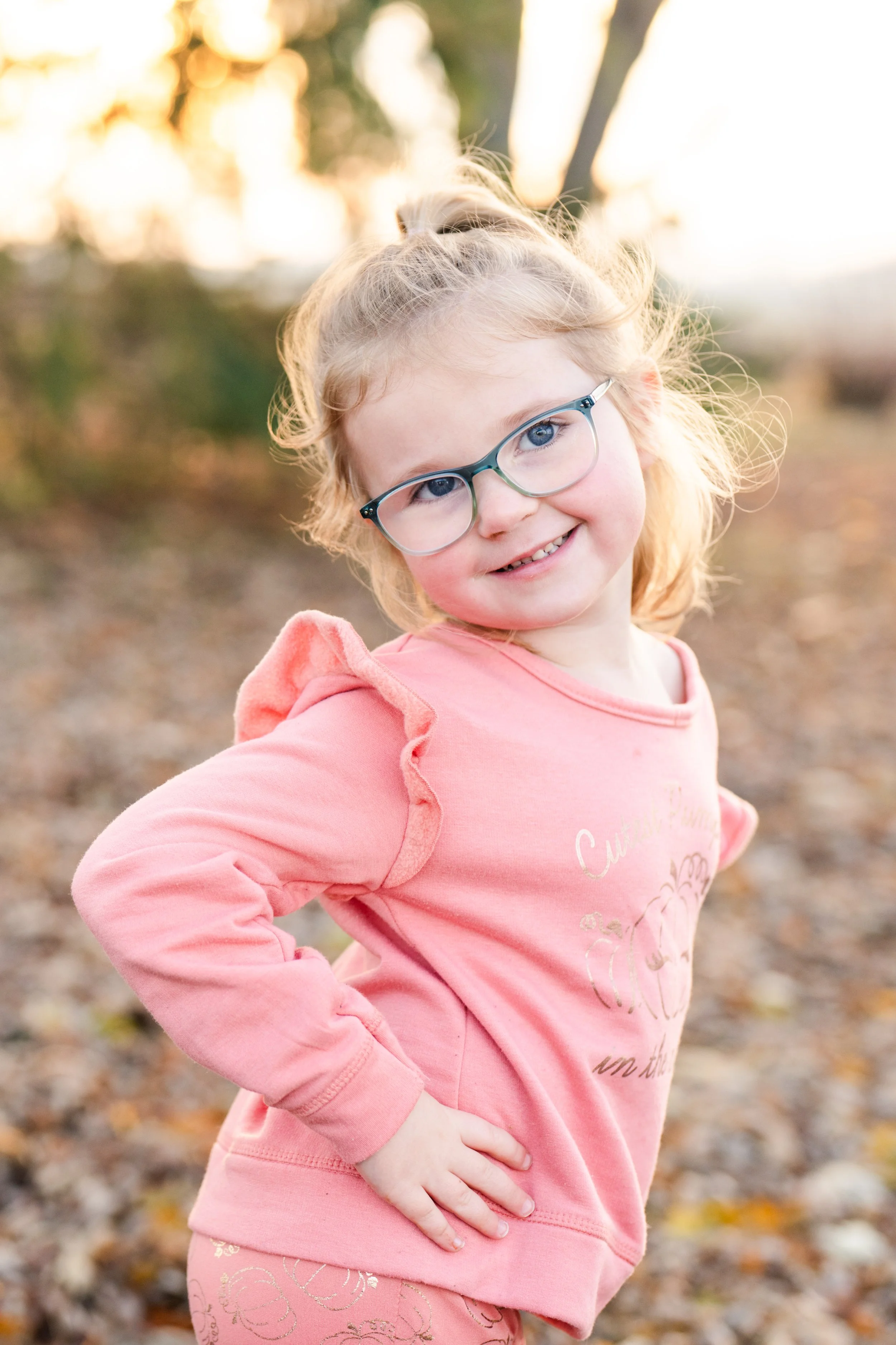 A young girl with blonde hair, glasses, and a pink sweatshirt with ruffled sleeves, smiling outdoors during sunset, taken by MN photographer LKW Photography.