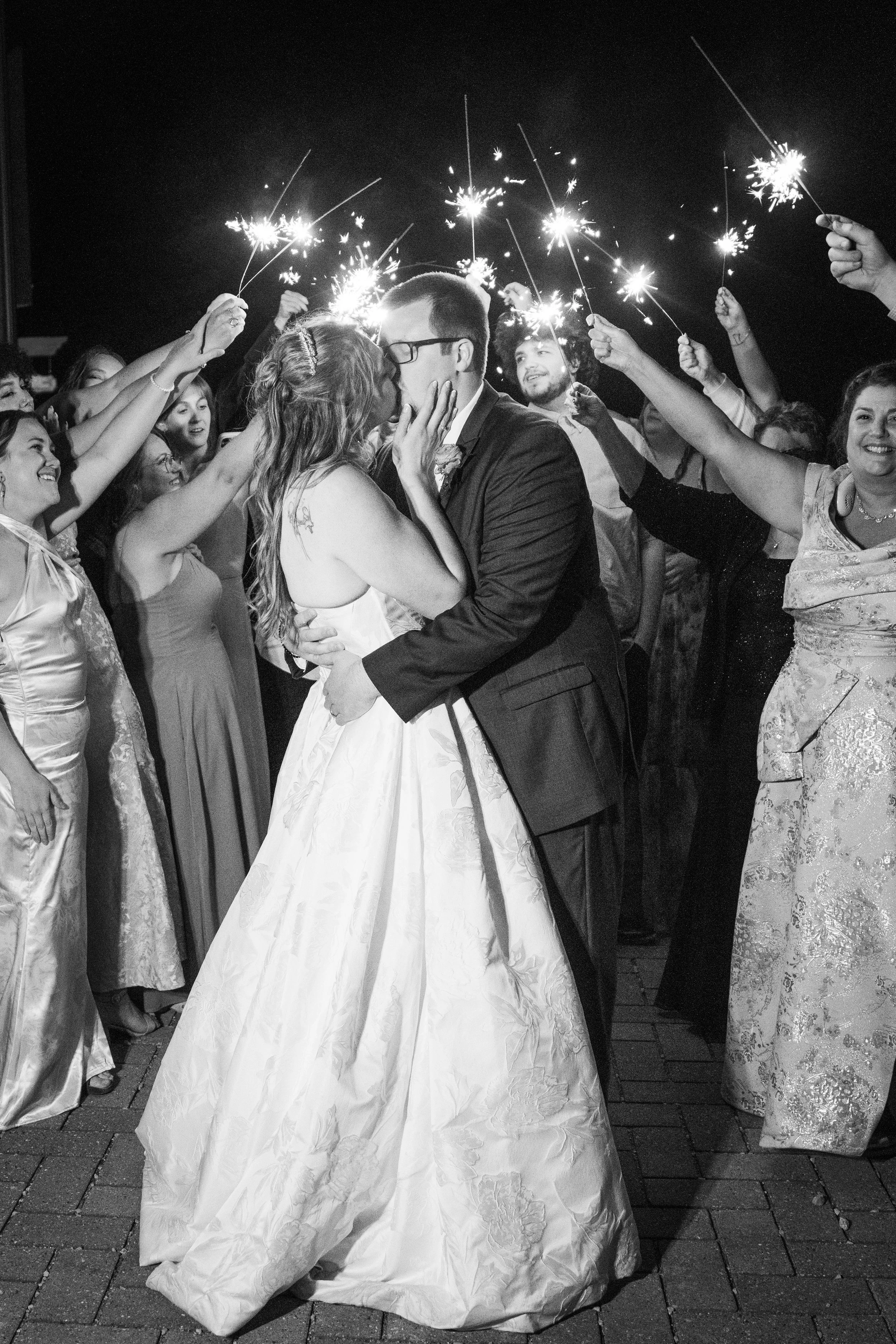 A bride and groom kissing surrounded by people holding sparklers at night; black and white photograph.