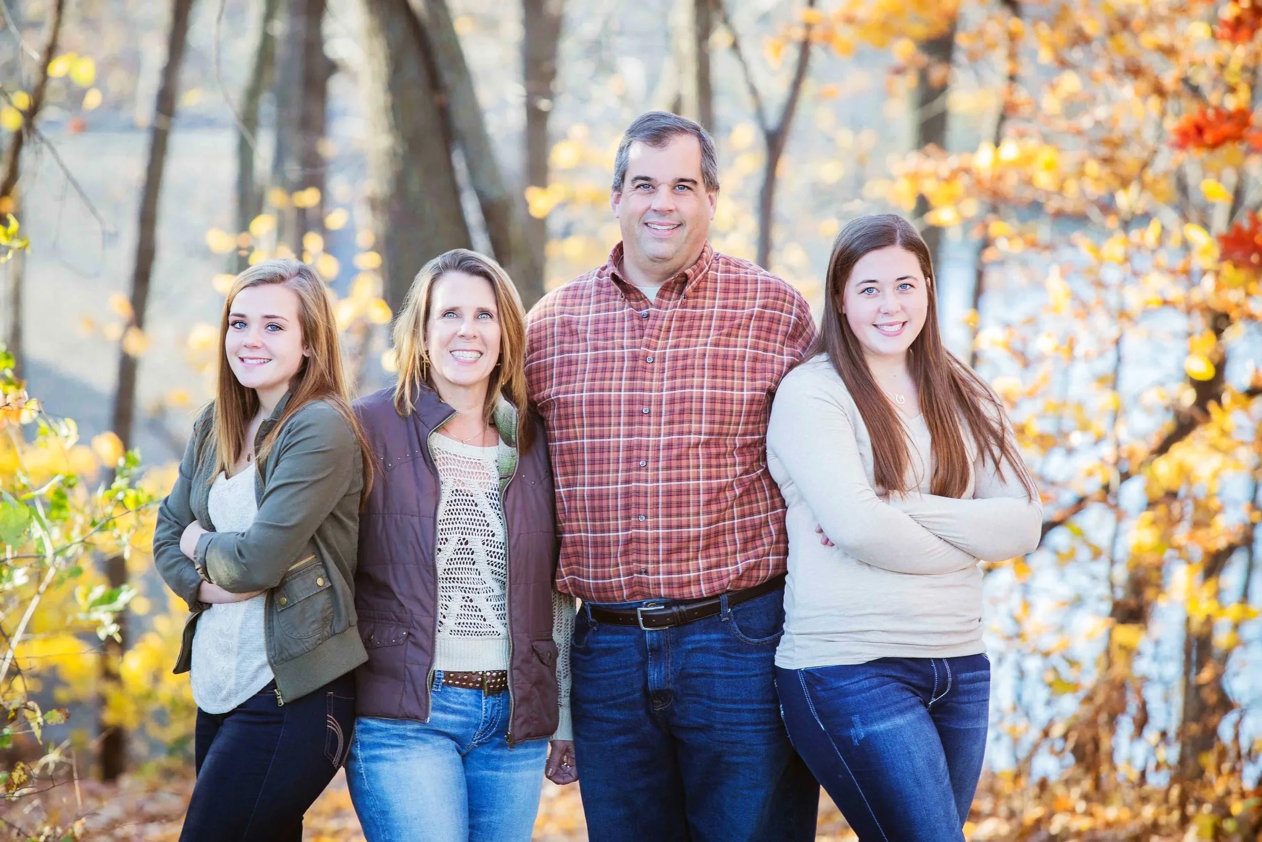 A family of five standing outdoors in a wooded area with fall foliage, smiling at the camera, , taken by Minnesota Family Photographer LKW Photography.