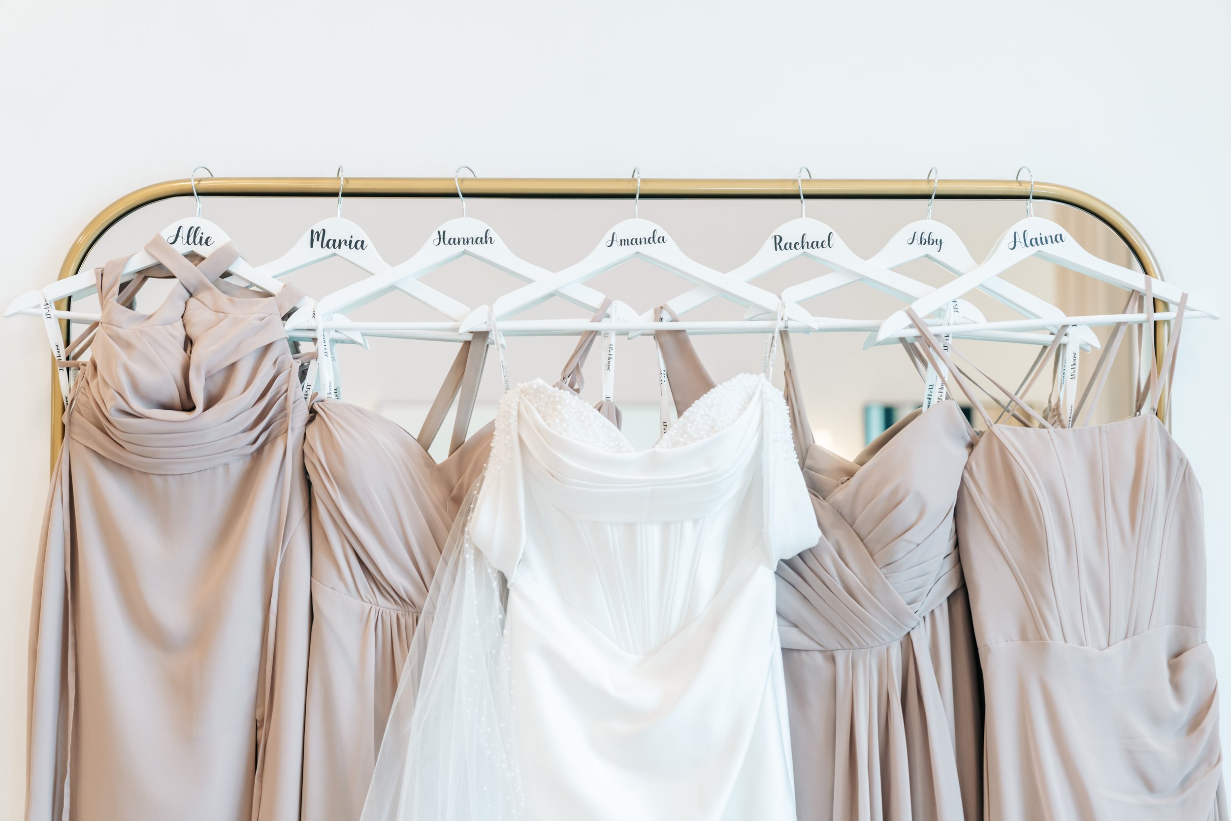 A clothing rack displaying dresses with name tags, including Allie, Maria, Hannah, Amanda, Rachael, Abby, and Alaina, with beige and white dress options.