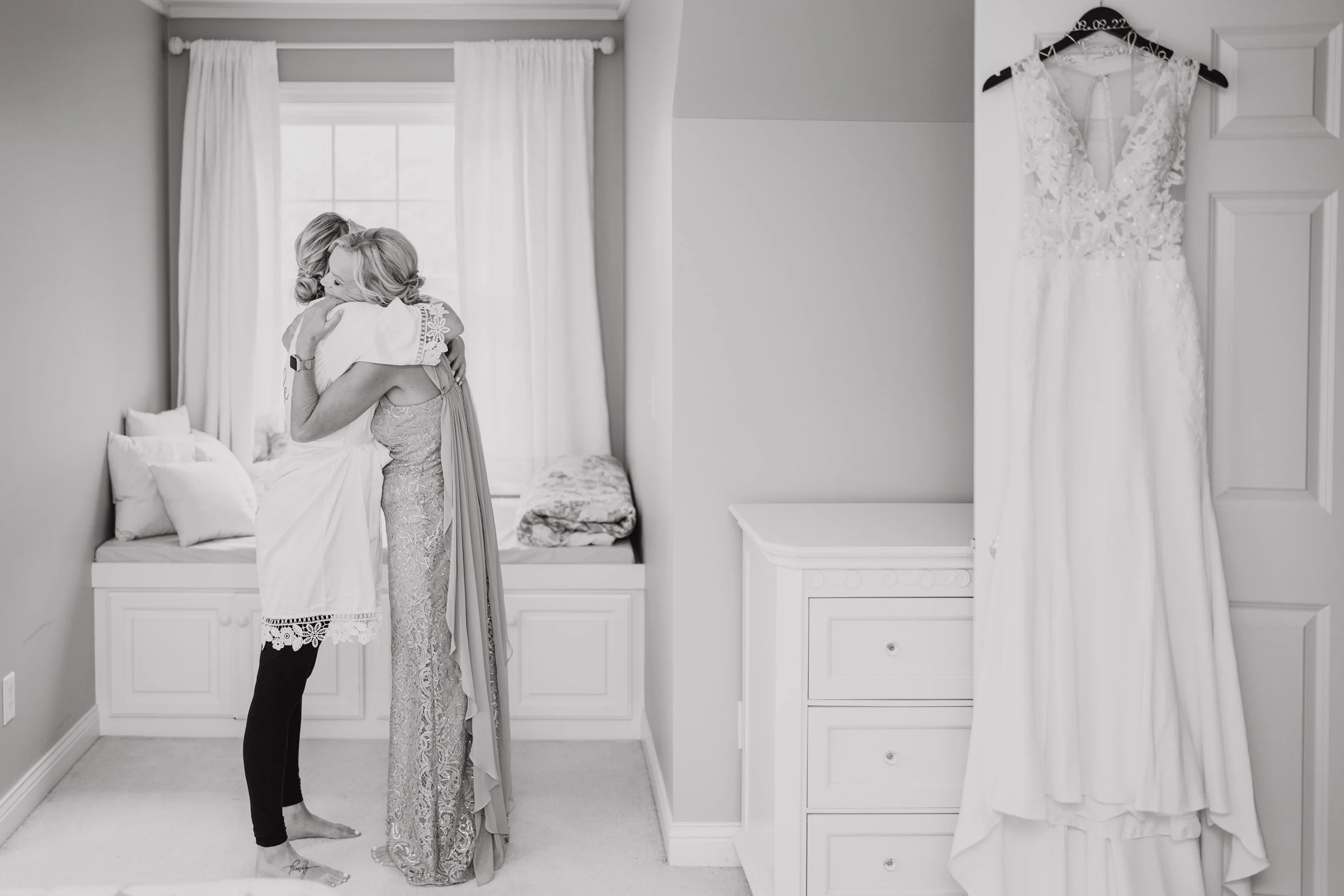 Two women hugging in a bedroom, with a white wedding dress hanging on a door on the right side of the image.