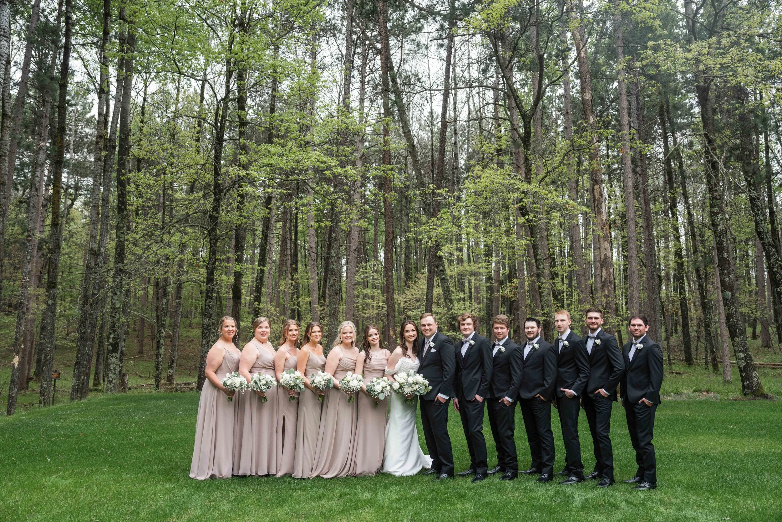 A wedding party standing in a forest, with the bride and groom in the center and bridesmaids and groomsmen on either side, all dressed in formal attire.