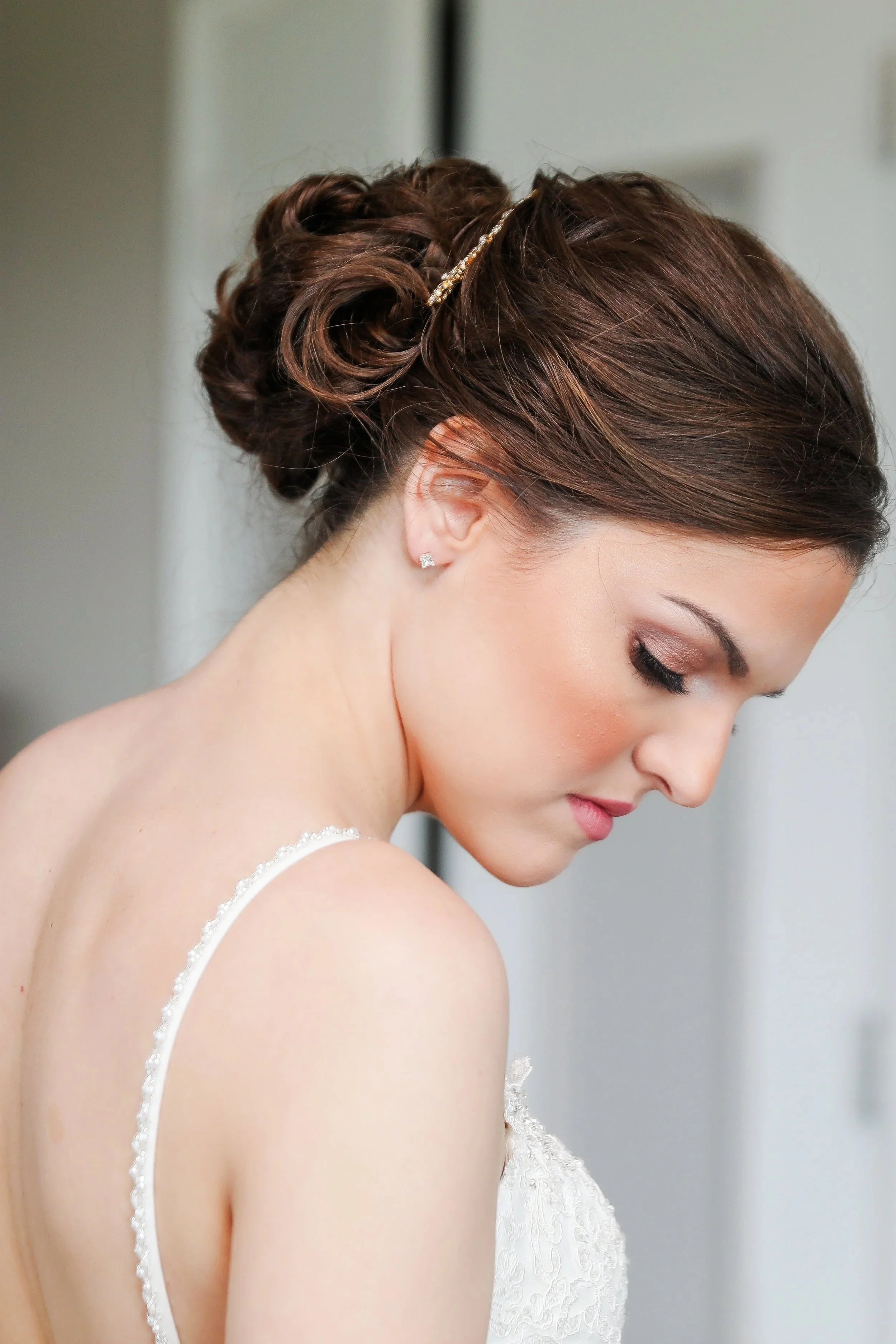 Close-up of a bride with styled updo hair, wearing a white lace wedding dress and diamond stud earrings, looking down with a serene expression.
