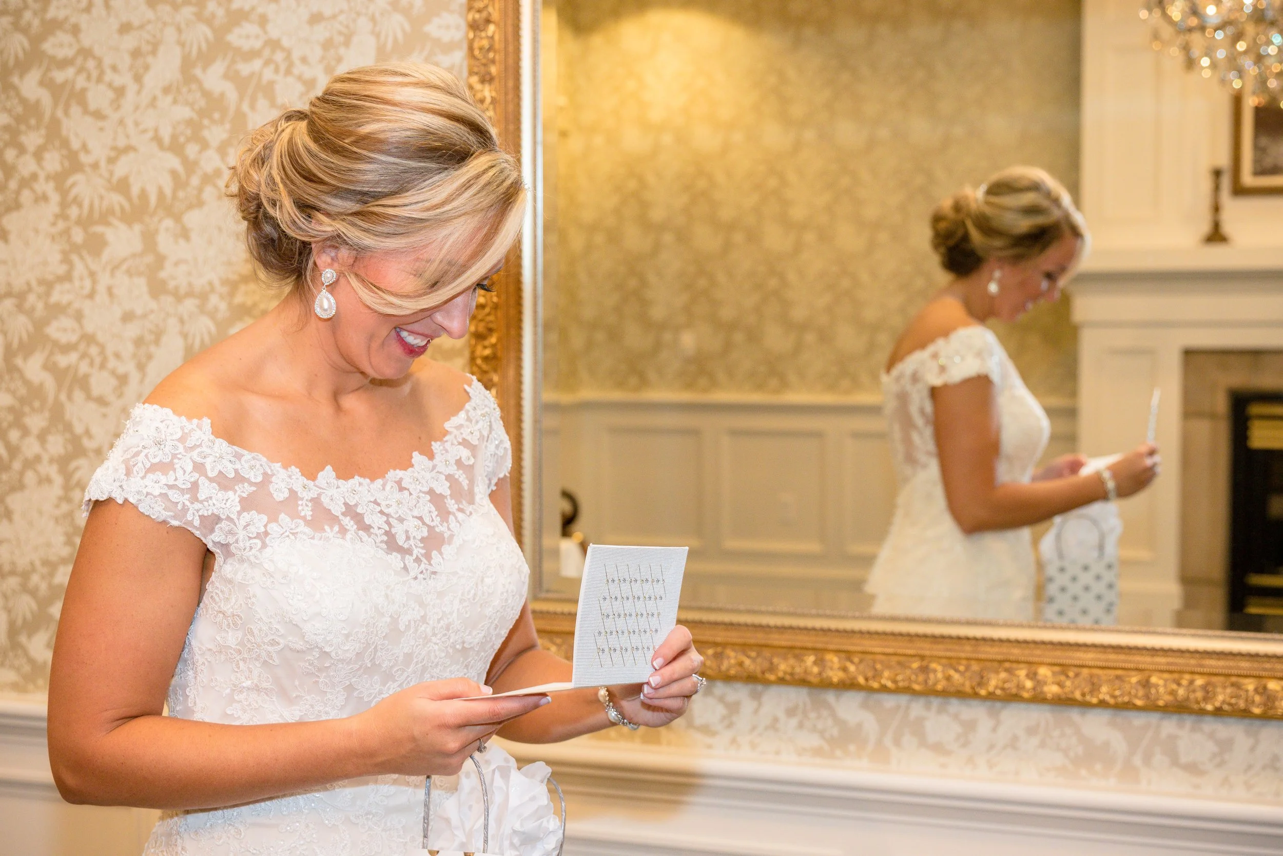 A woman in a white lace wedding dress is smiling while looking at a card or note in her hand. She is in front of a large, ornate mirror, and her reflection shows her holding a similar item. The room has elegant wallpaper with a floral pattern and a f