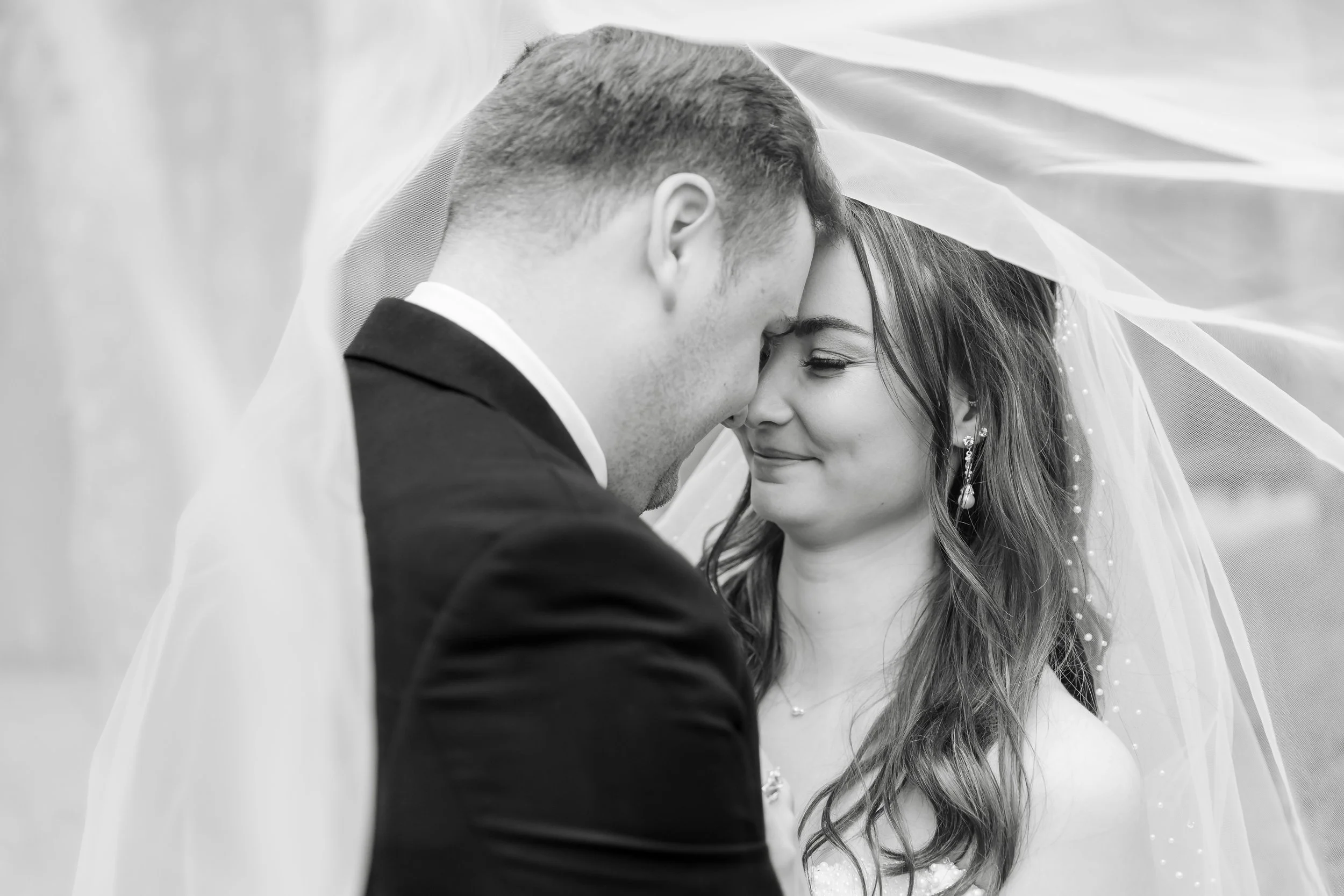Black and white photo of a groom and bride with foreheads touching under a sheer fabric veil during a wedding ceremony.