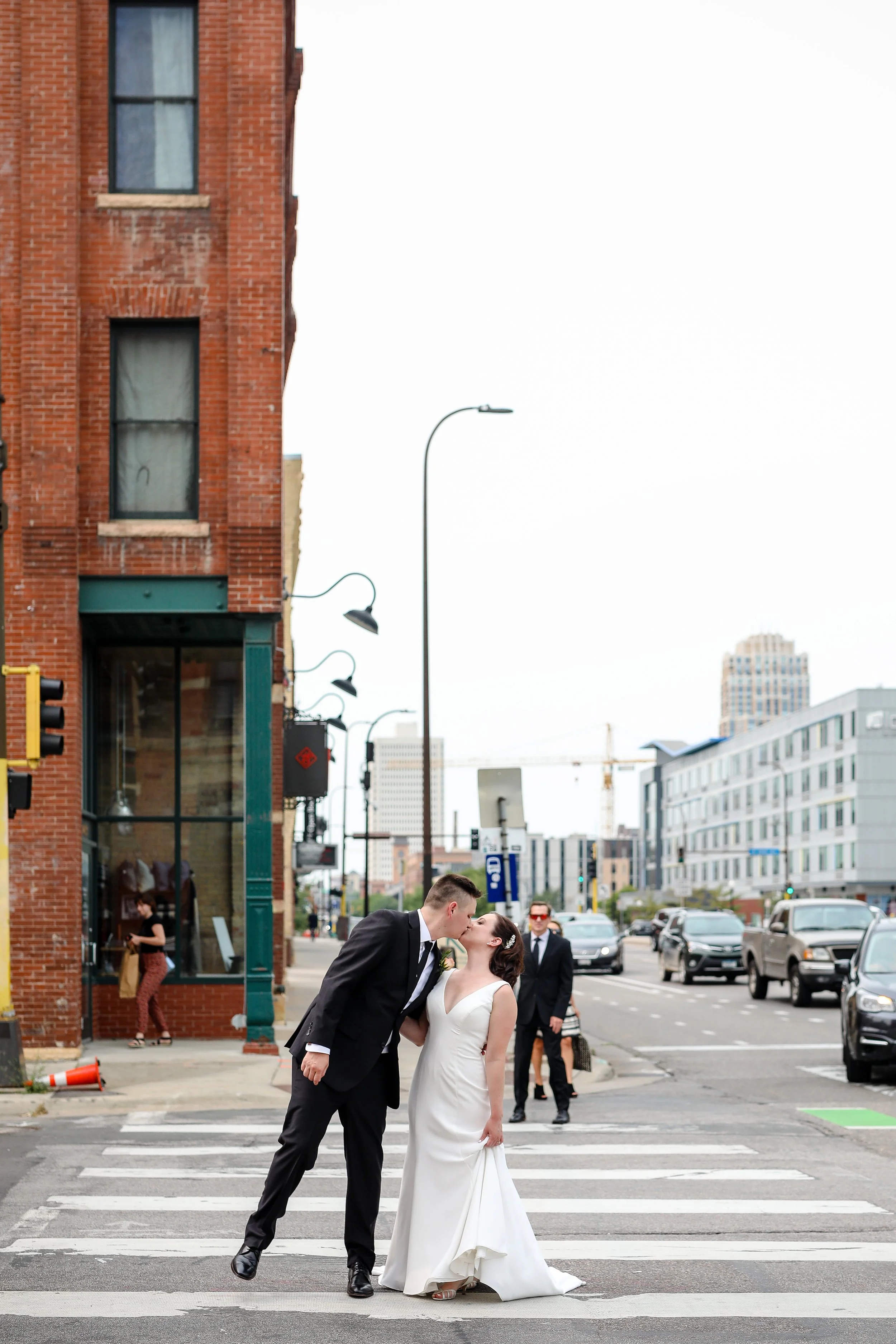 A newlywed couple sharing a kiss while standing in the crosswalk on a city street, with pedestrians and cars in the background.