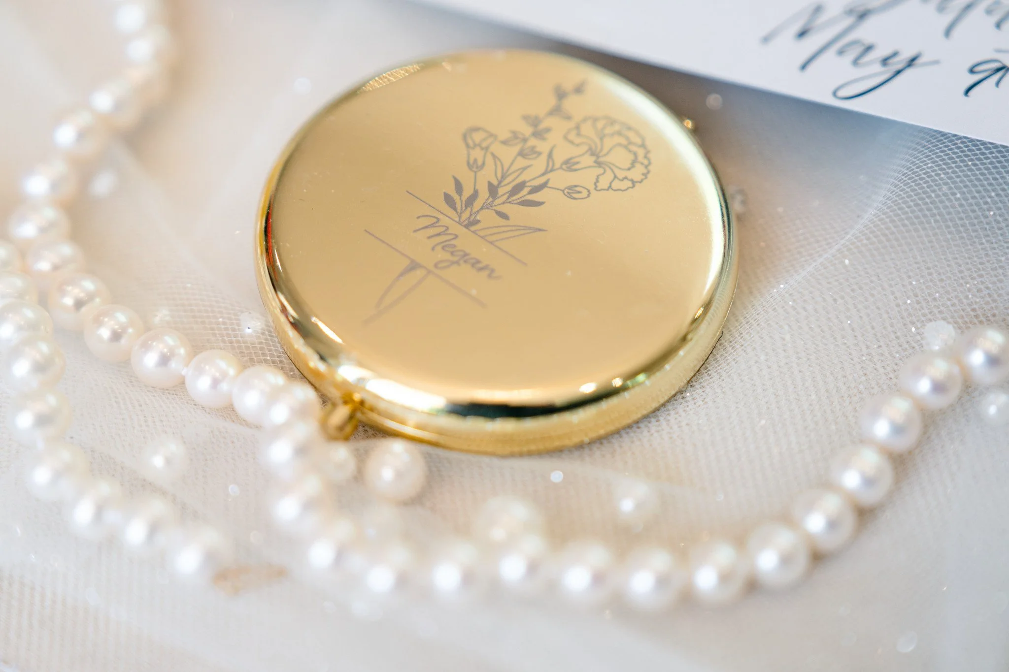Wedding details with an antique gold pocket mirror with the name Megan inscribed on it, laying on a veil with pearls, and surrounded by a pearl necklace and bracelet.