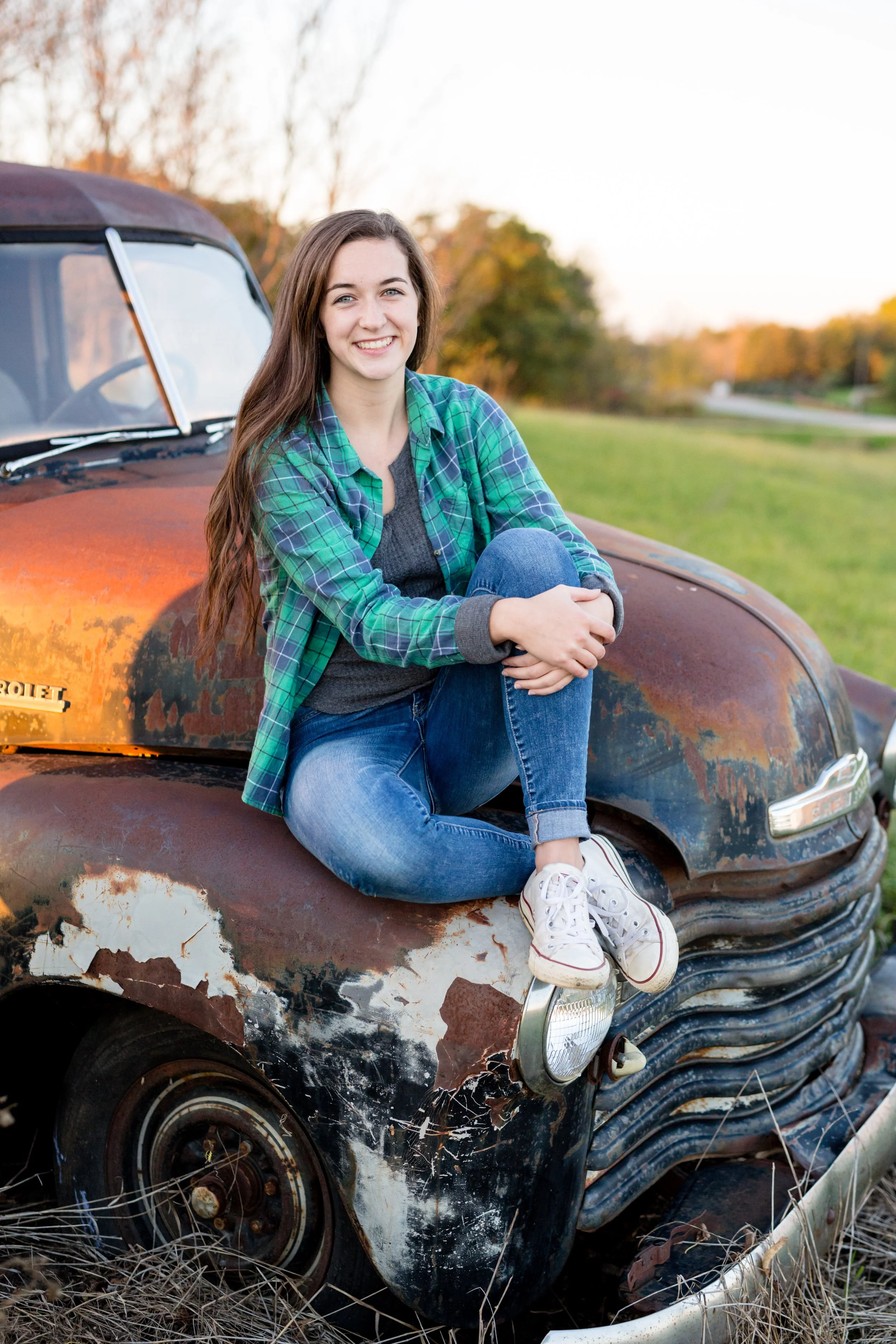 A young woman in jeans, a gray shirt, and a green checkered flannel shirt sitting on the hood of an old, rusted Chevrolet truck outdoors in a rural area during sunset, taken by Minnesota Senior Photographer, LKW Photography.