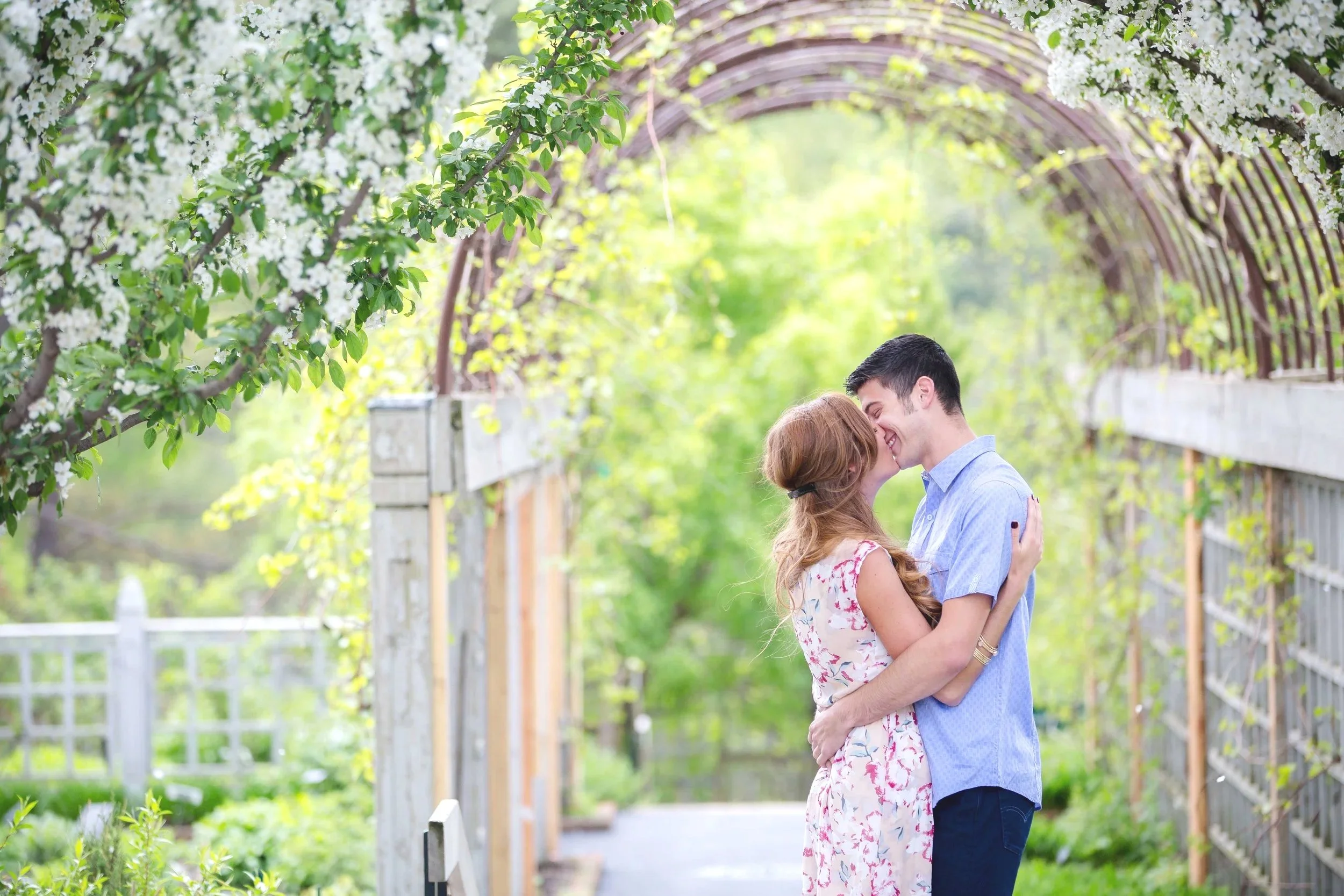 A couple sharing a kiss on a garden pathway under a wooden archway covered with white blossoms, surrounded by lush green foliage, taken by Minnesota Wedding Photographer LKW Photography.