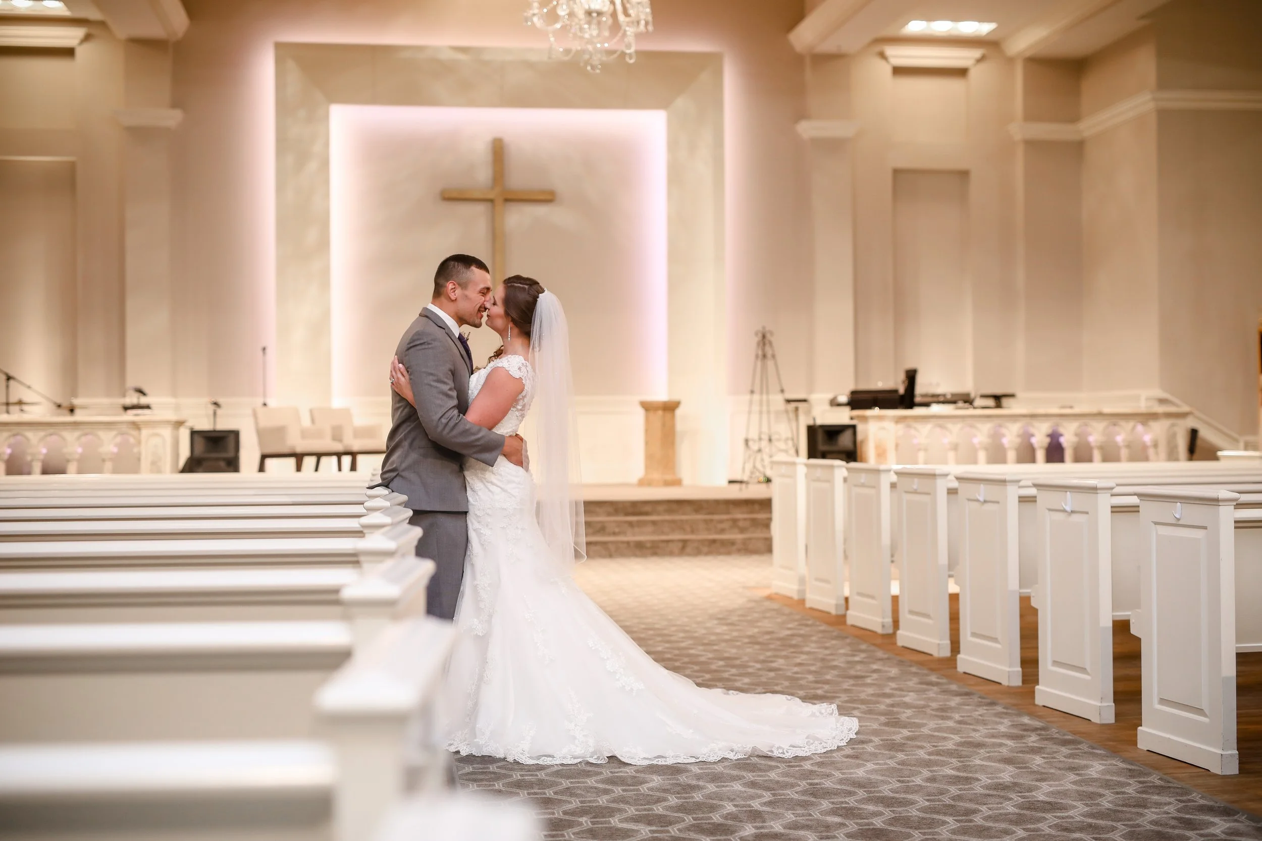 A bride and groom embracing and about to kiss inside a church, with pews, a cross on the wall, and a decorated altar visible in the background.