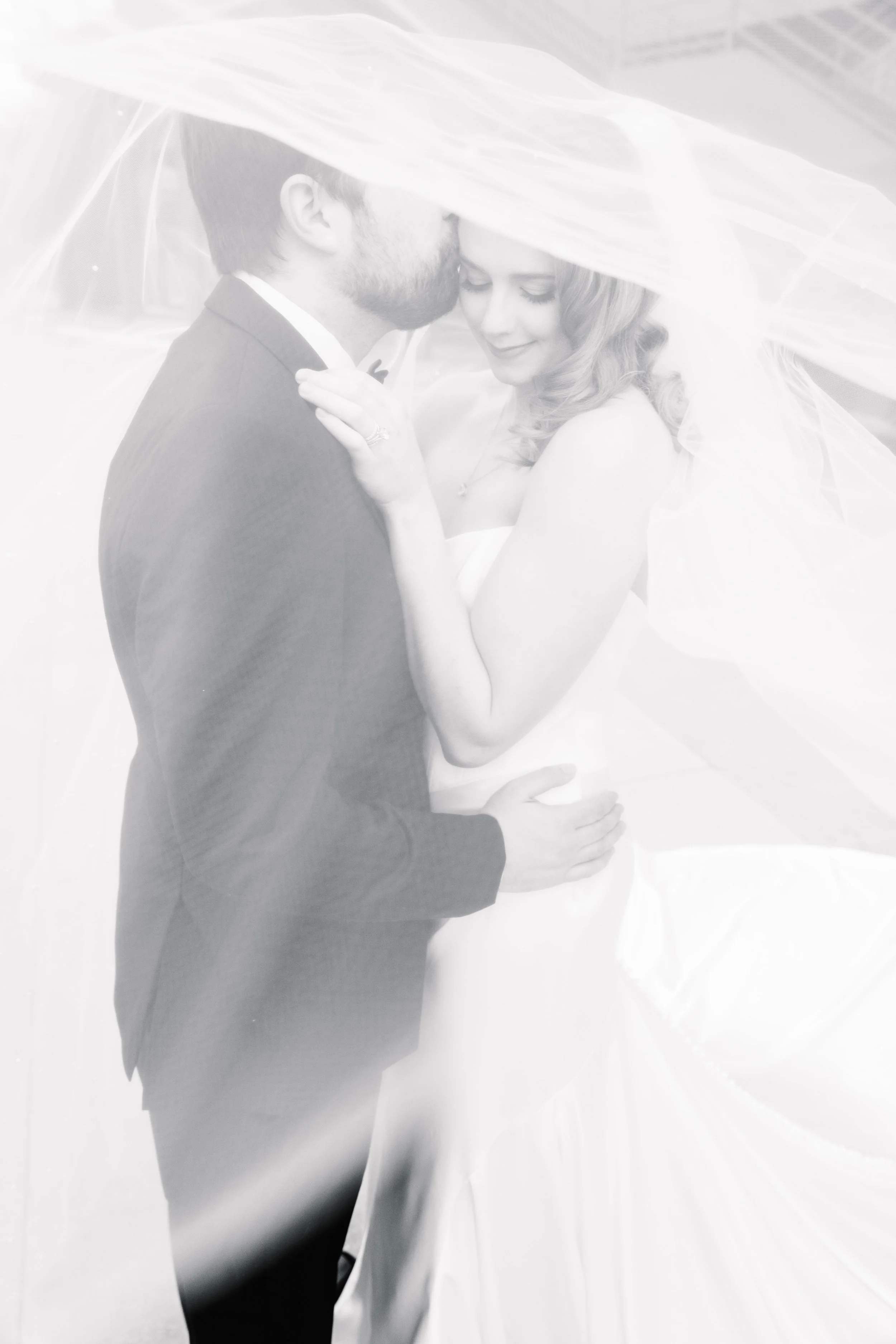 A black-and-white photo of a couple on their wedding day, embracing beneath a sheer veil. The man, in a suit, is kissing the woman's forehead, who smiles with her eyes closed, wearing a wedding gown and an engagement ring.