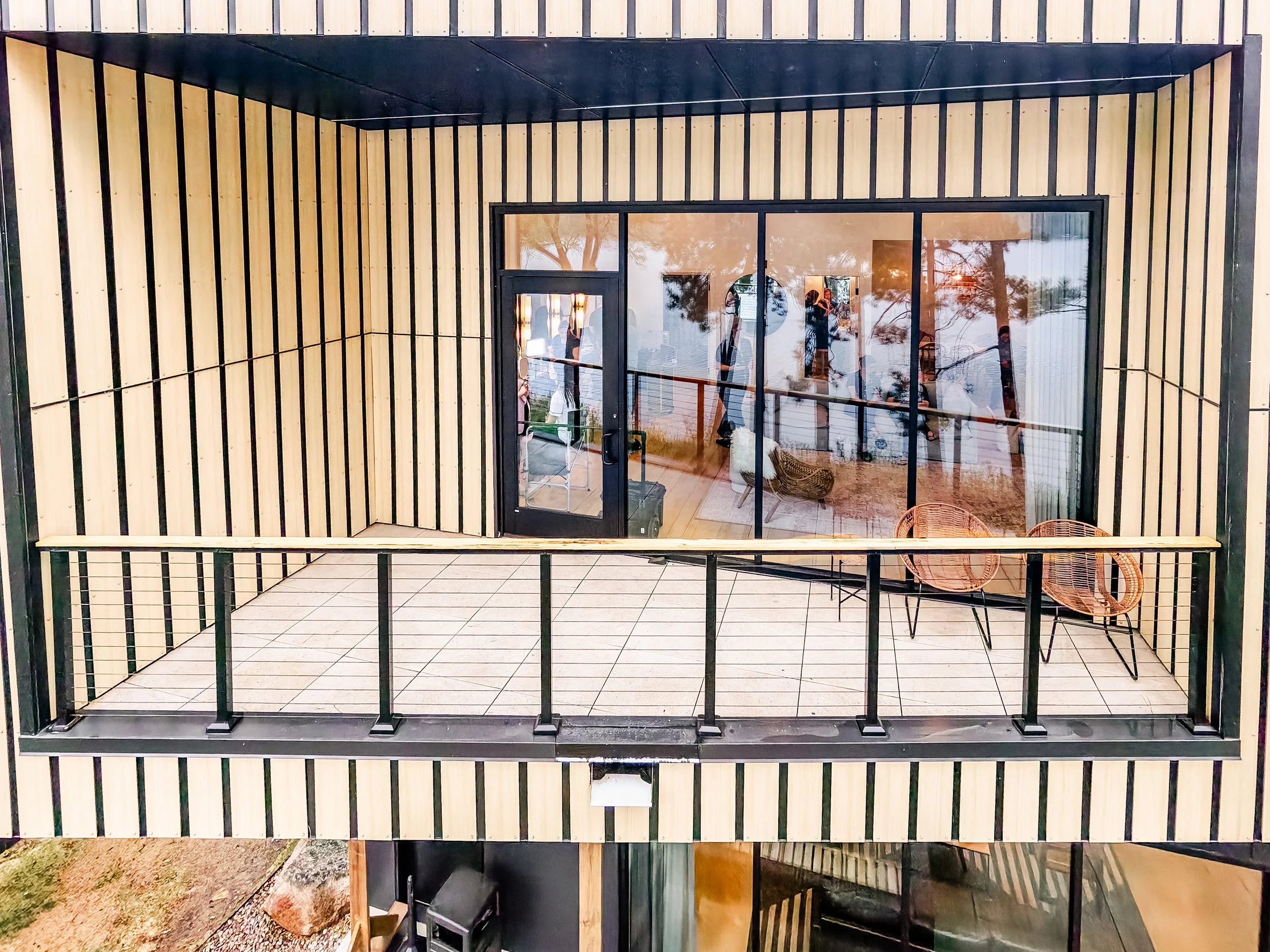 The exterior of a modern building with vertical wooden slats and large glass doors, reflecting trees and a person inside. There are two wicker chairs outside and a tiled floor.