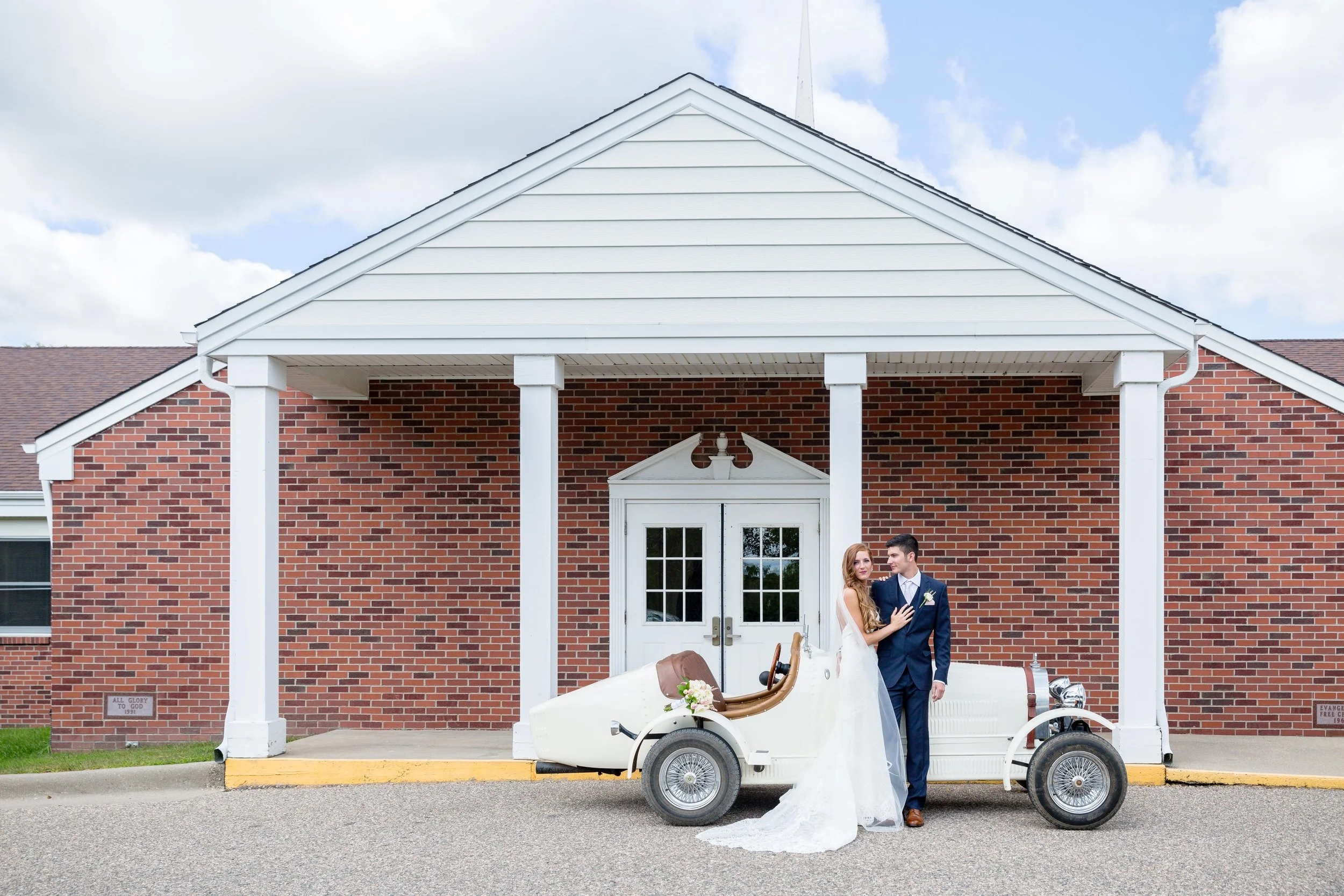 A bride and groom stand next to a vintage white car with brown leather seats, parked in front of a brick church, photographed by Minnesota based wedding photographer, LKW Photography.