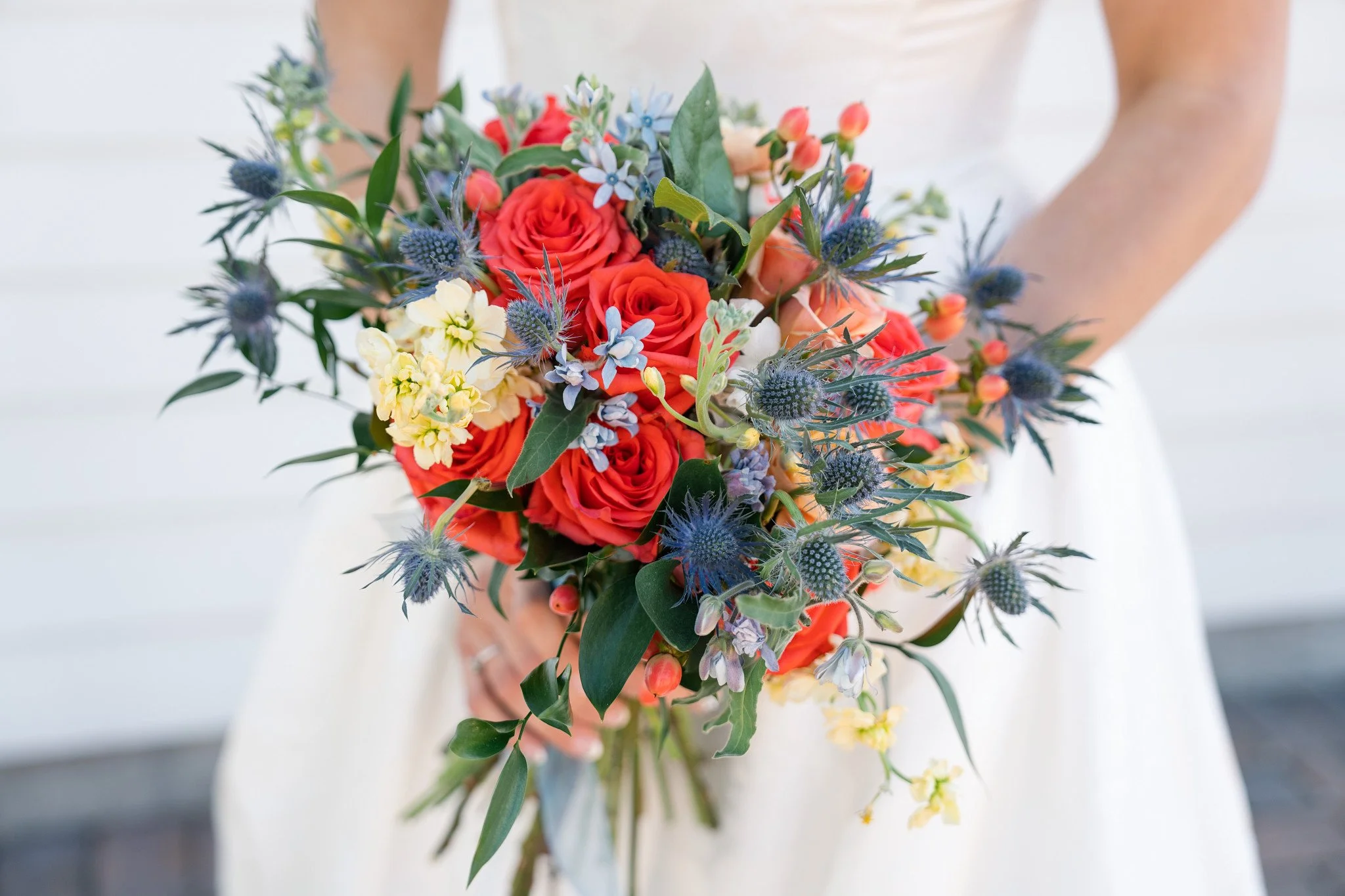 A bride holding her wedding bouquet of mixed flowers including red roses, blue thistles, yellow flowers, and small pink buds.