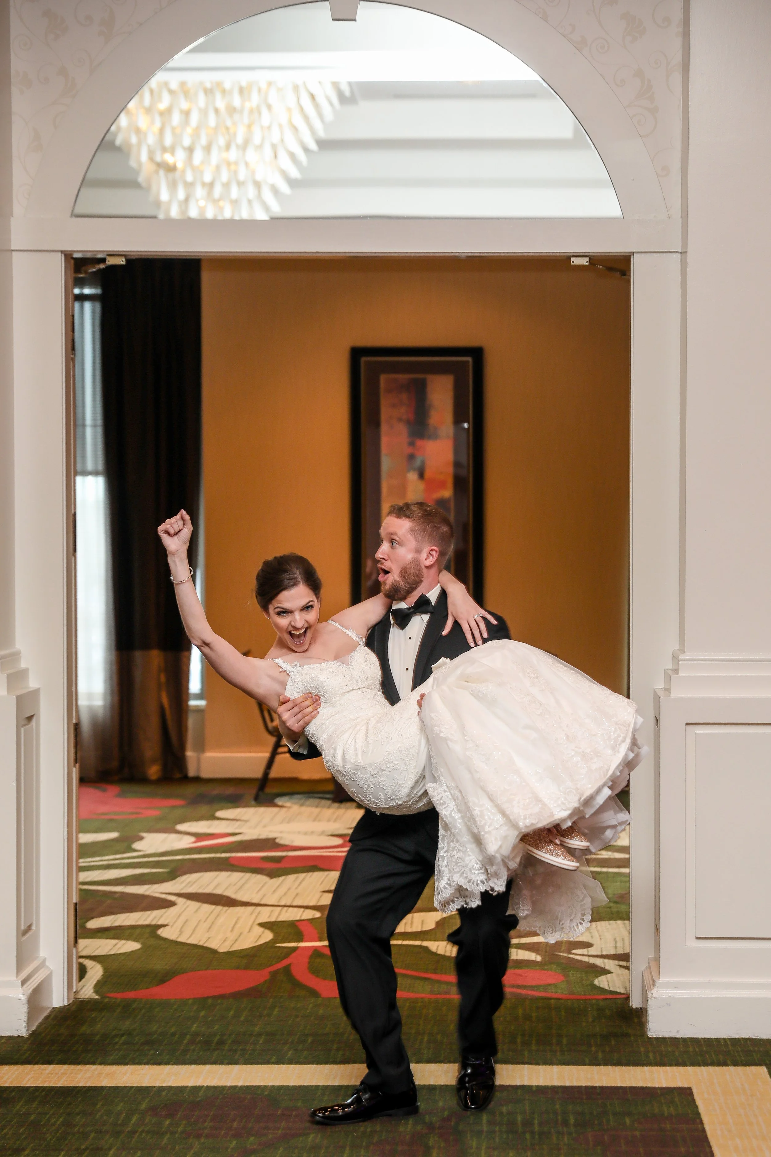 A man in a tuxedo lifts a woman in a white wedding dress, cheering with raised arm, inside an elegant room with colorful carpet and framed artwork.