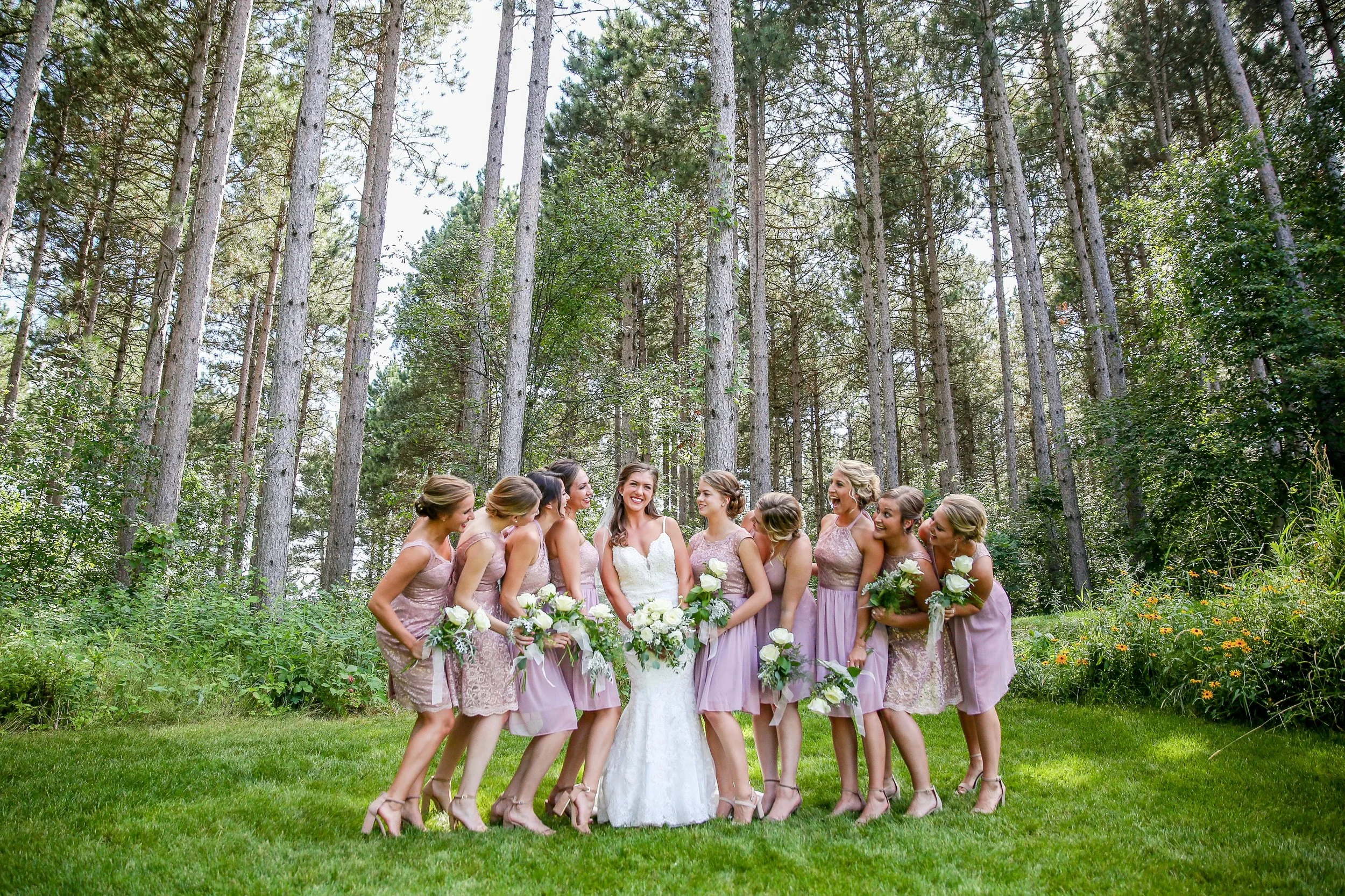 A bride and nine bridesmaids posing outdoors in a green, wooded area during a wedding celebration, photographed by Minnesota based wedding photographer, LKW Photography.