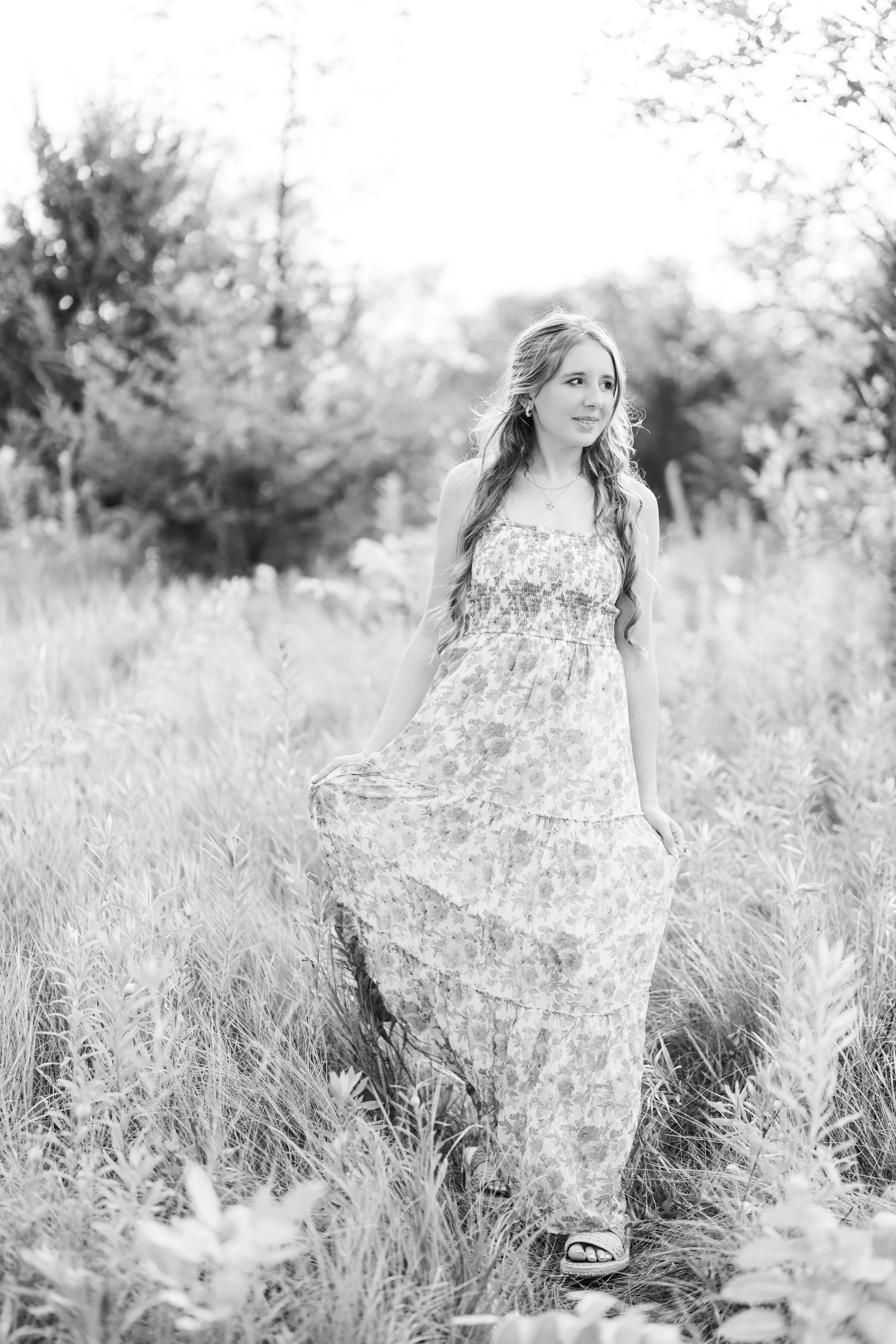 Minnesota senior photographer, LKW Photography, photographs a high school senior in a floral maxi dress walking through a grassy field, holding the sides of her dress, with trees in the background.