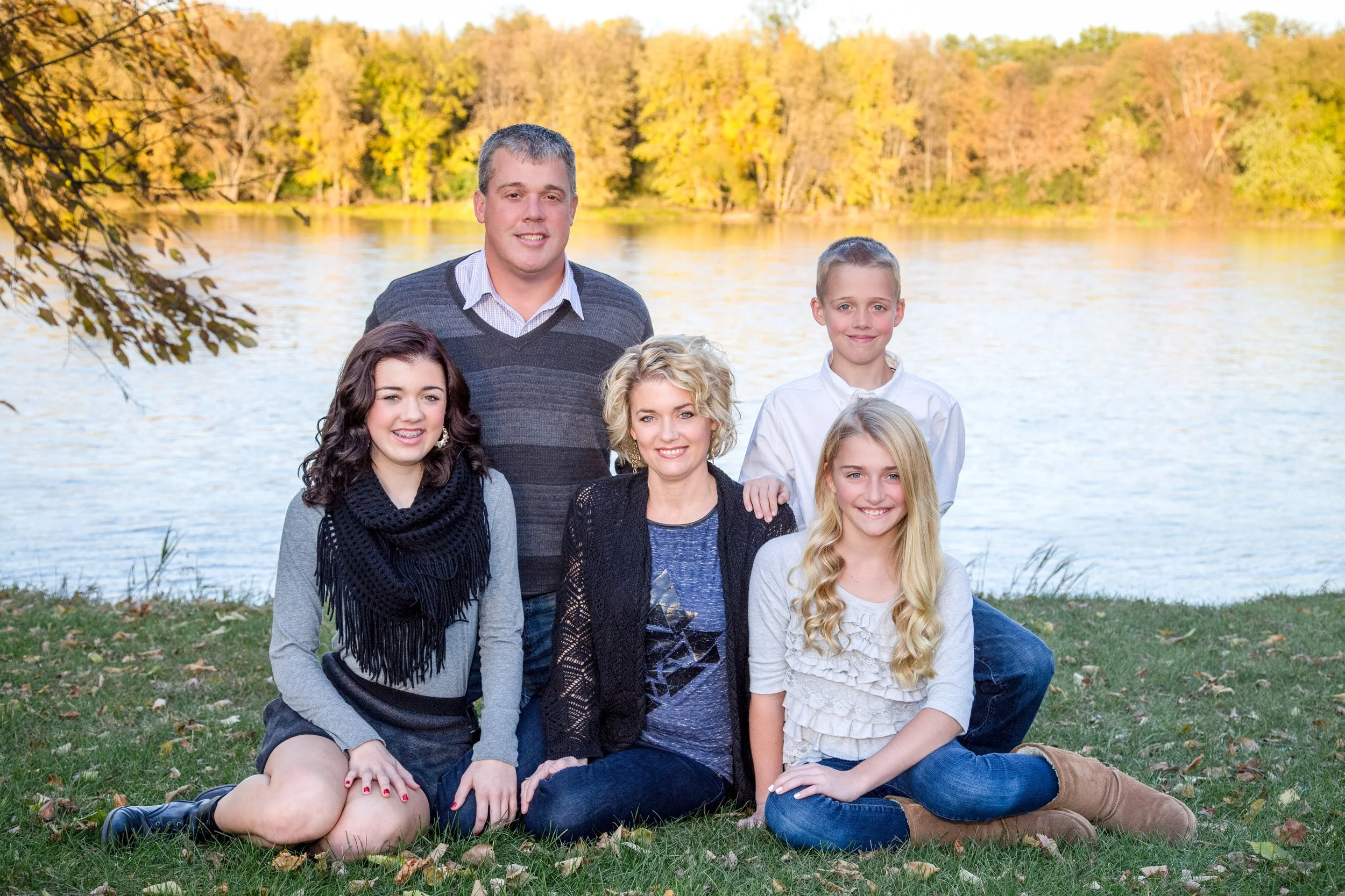 Family of six sitting on grass near a lake with trees in fall colors in the background, smiling at the camera, taken by Minnesota Portrait Photographer LKW Photography.